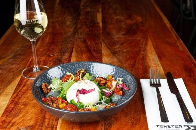Een salade met burrata kaas, cherrytomaten, rucola en basilicum op een donkere, ronde kom, naast een glas witte wijn en een vork en mes op een witte servet op een houten tafel.