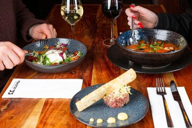 Twee mensen dineren met salades en een gerecht met vis of garnalen, wijn en water op een houten tafel met bestek en servetten.