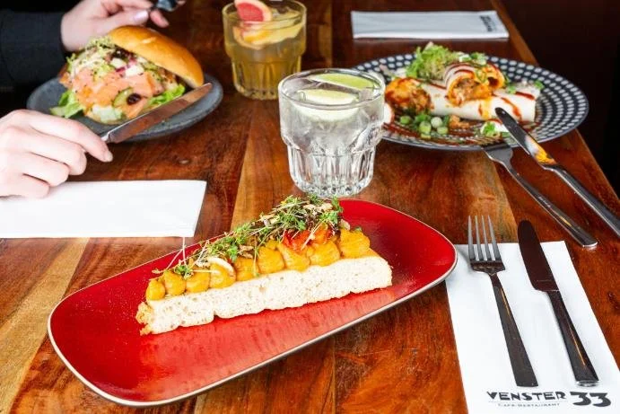 Gezellig diner met diverse gerechten op een houten tafel, inclusief een plank met pastinaak, een glas water, een gerecht met sushi, en een sandwich.