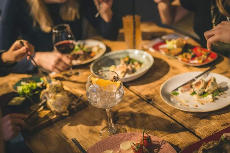 Gezelschap dineert aan een houten tafel met diverse gerechten en drankjes, waaronder een gin-tonic met sinaasappel en een glas rode wijn.