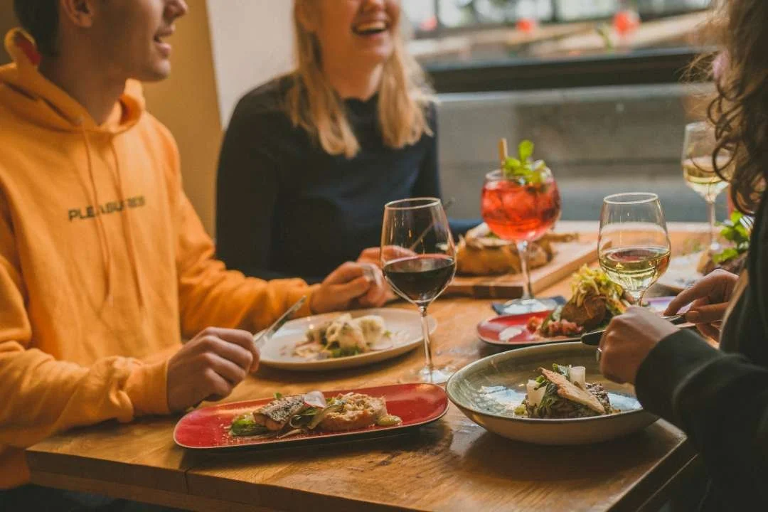 Vier mensen zitten aan een tafel in een restaurant, eten en drinken genieten van een maaltijd met wijn en cocktails.
