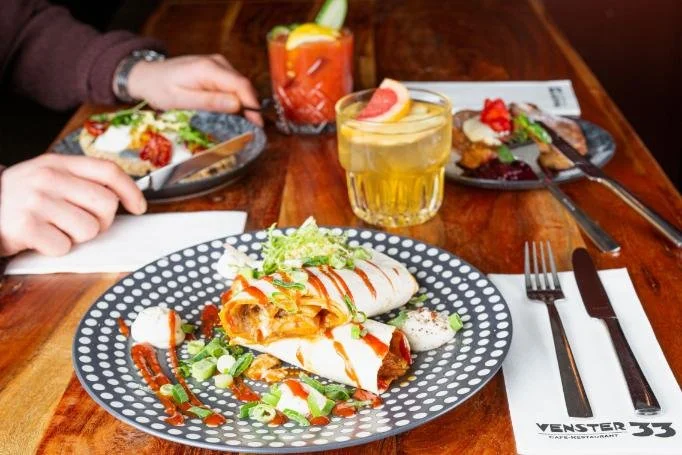 Gereserveerde tafel met drie borden gevuld met Mexicaanse enchiladas, limoengarnituur, en twee cocktails, in een restaurant.