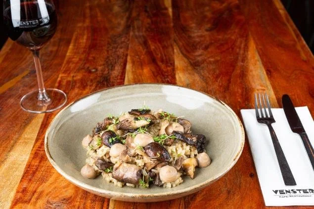 Een bord met een gerecht van kikkererwten, aubergine en champignons op een houten tafel. Er staat ook een glas rode wijn en bestek met een wit napkin naast het bord.