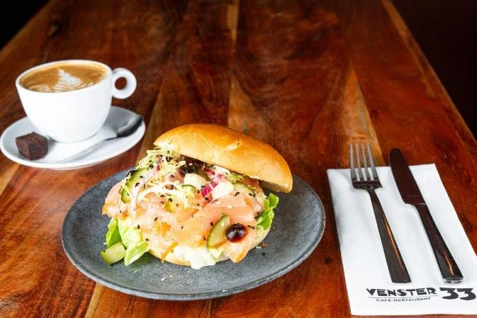 Sandwich met zalm en groenten op een donkere bord, koffie met melk en schuim, en een kopje thee met suiker op een houten tafel.