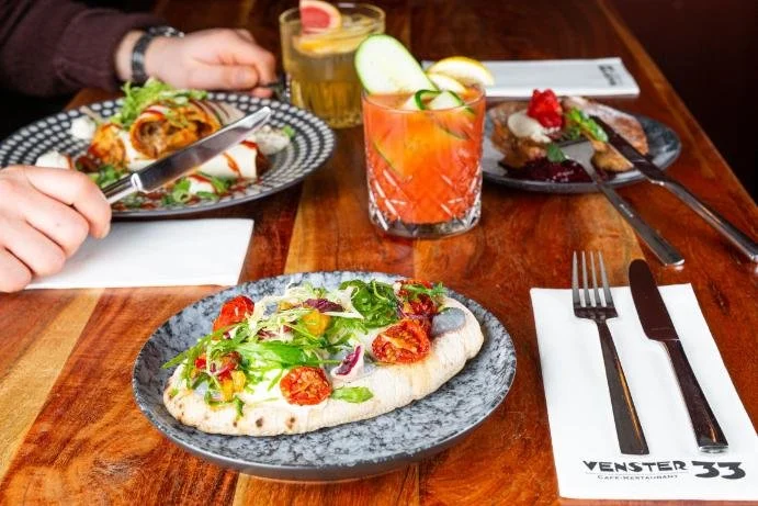 Een tafel met verschillende gerechten en dranken, waaronder een pizza met sla en tomaten, een glas met een roodgekleurde cocktail met citrus en komkommer, een gerecht met wat gefrituurde snacks, en een glas met een gele drank. Bestek ligt naast een w