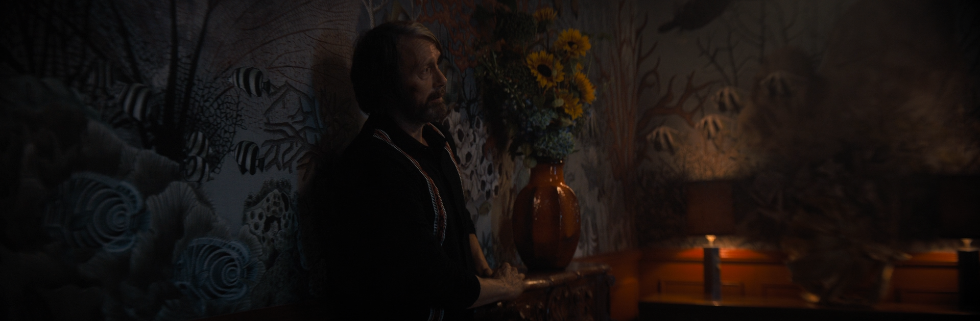 A man with dark hair and a beard sitting on a bench in a dimly lit room, with a large vase of flowers behind him and a small lamp illuminating the background.