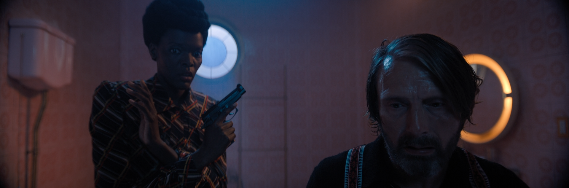 A scene from a movie showing a woman holding a gun with a concerned expression and a man with disheveled hair and a frightened look, in a dimly lit room with pink patterned wallpaper and circular wall decor.