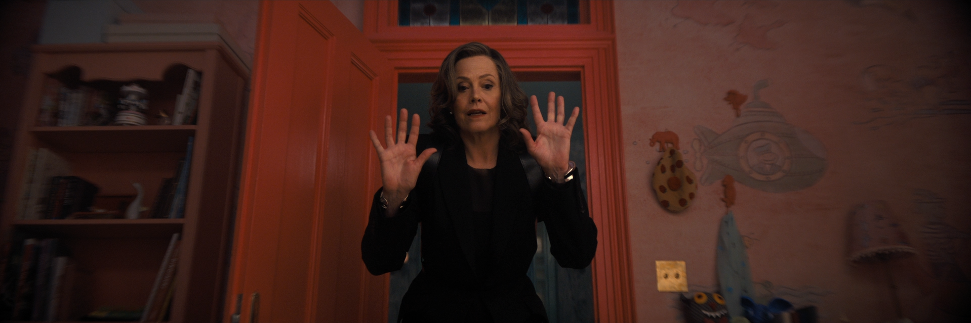 A woman with shoulder-length curly hair, dressed in black, is standing in a colorful room with pinkish walls and shelves filled with books and decorative items. She holds her hands up as if surprised or explaining something.