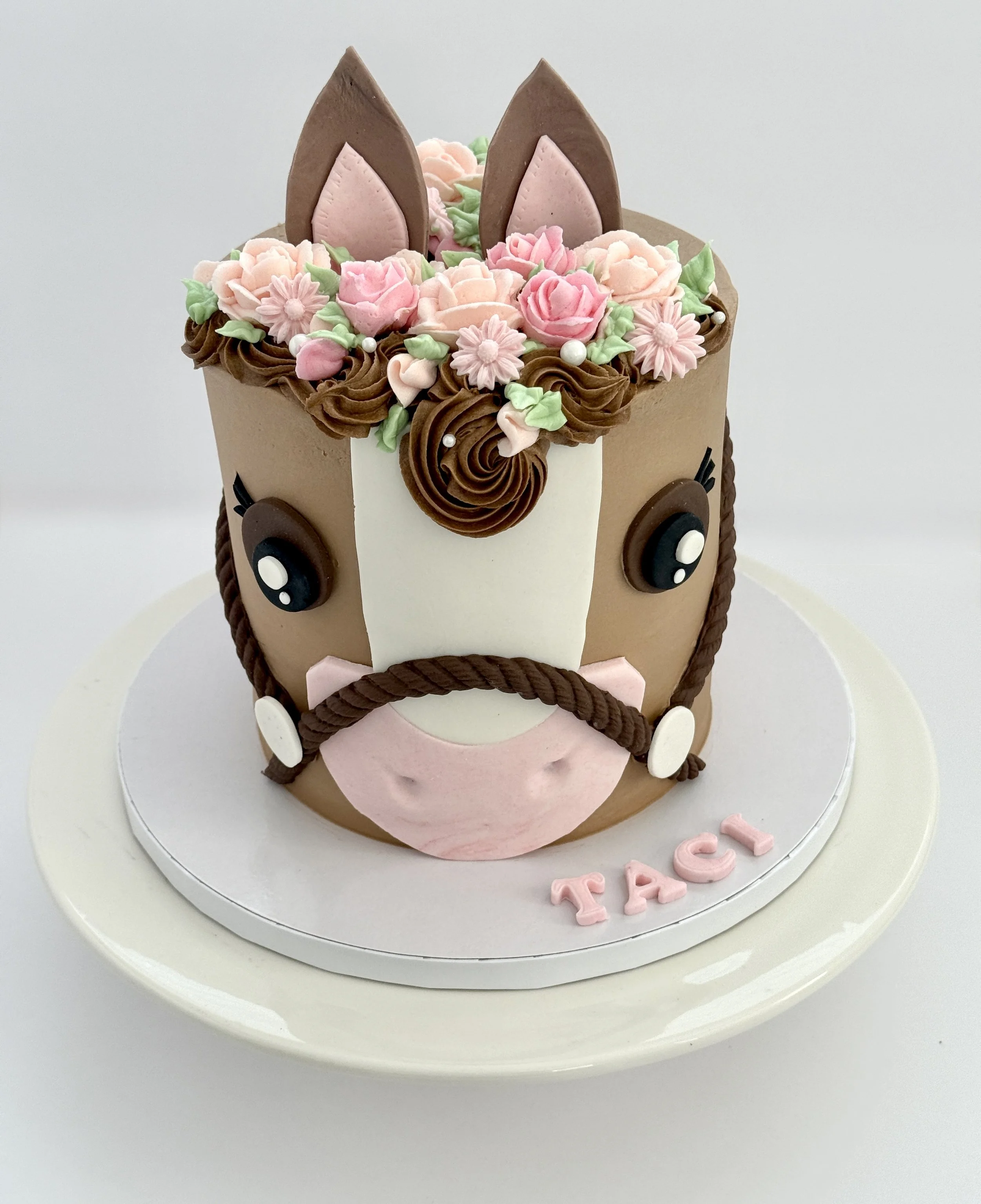 A cake decorated with pink and white floral icing, chocolate and pink cookie ears resembling a horse, and the name 'Fay' in pink letters on a white base.