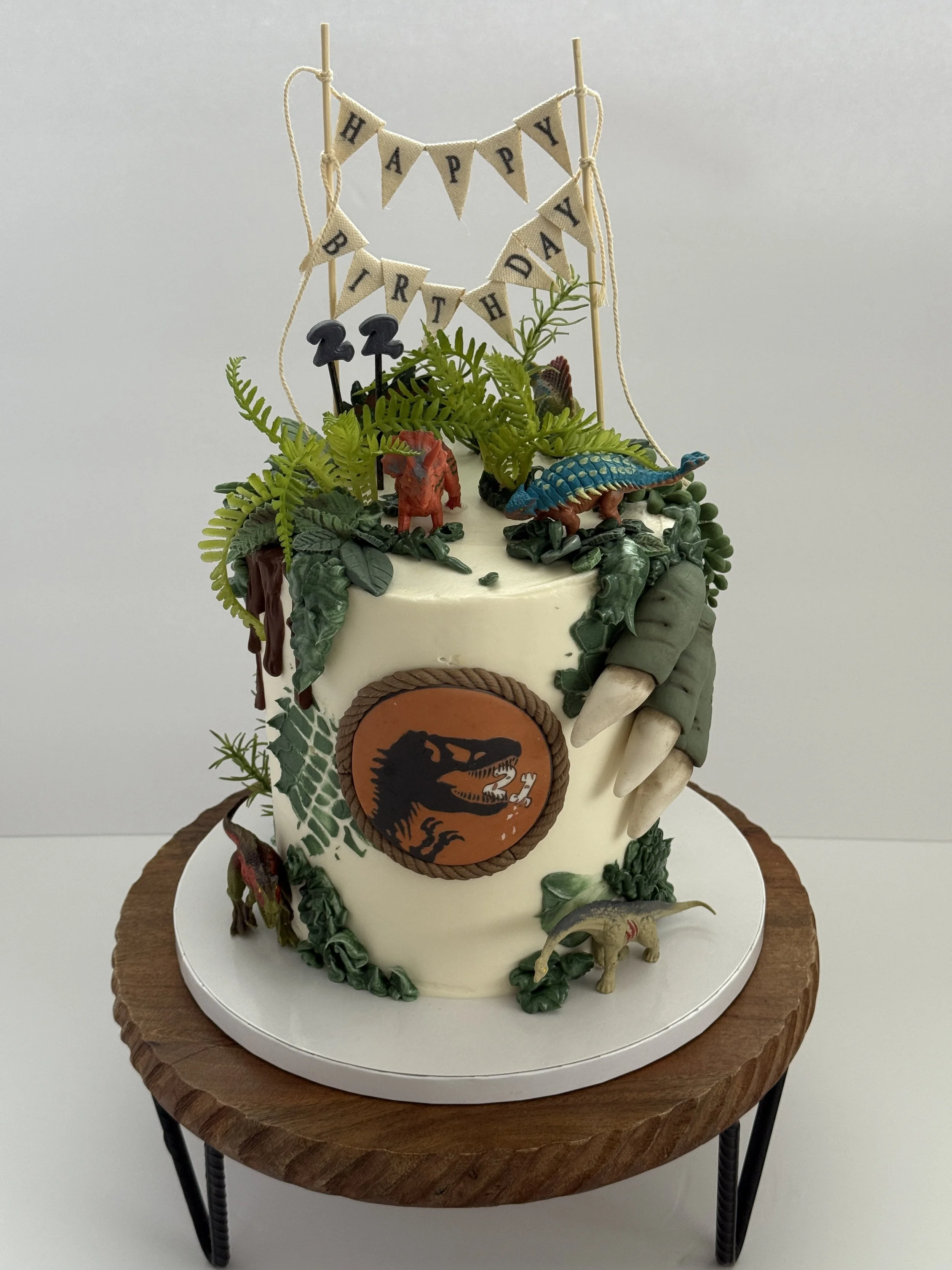 A multi-layered birthday cake decorated with dinosaur-themed figurines, green foliage, and a 'Happy Birthday' banner on top. The cake has a Jurassic Park logo on the side and is placed on a wooden table.