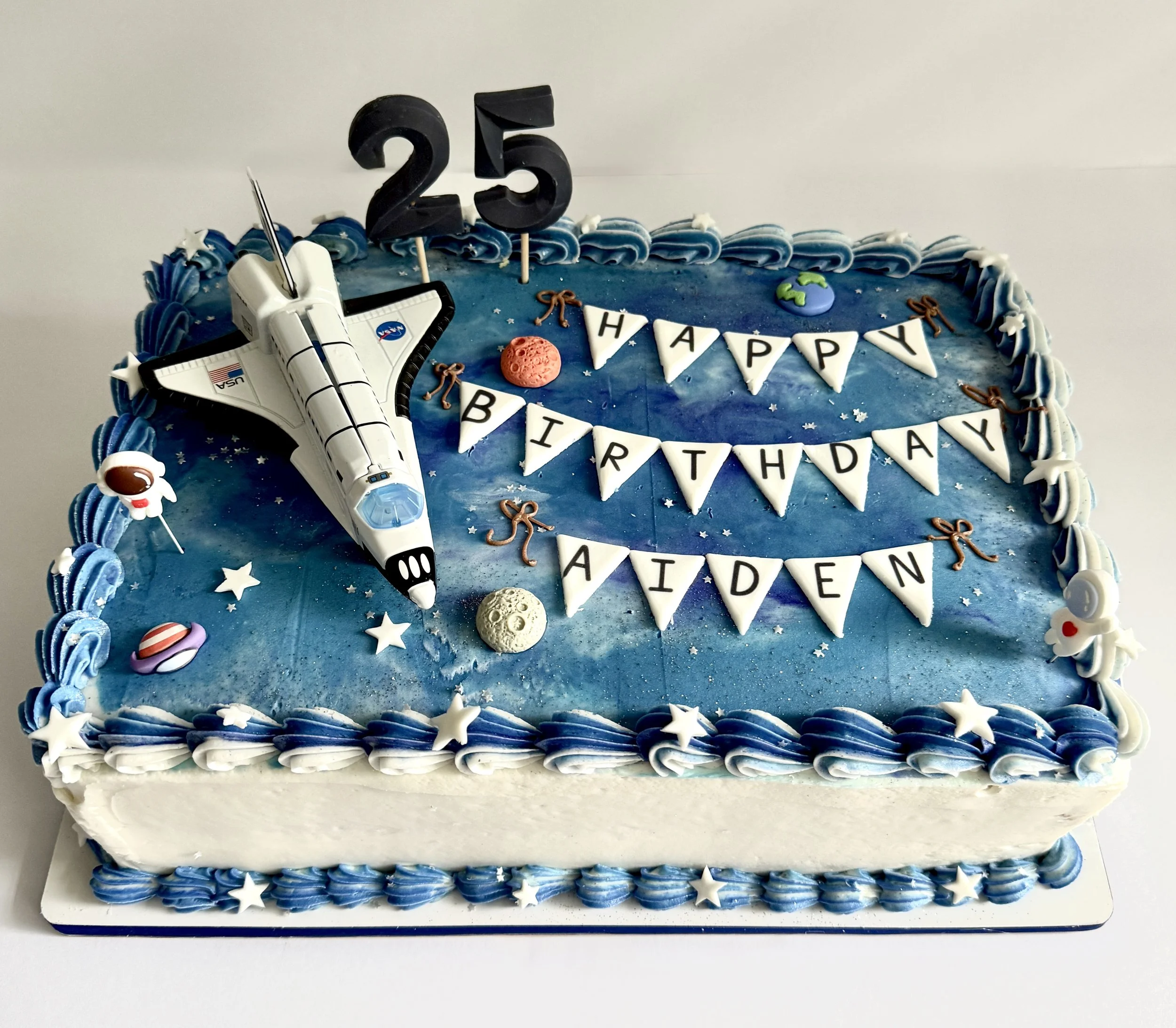 Space-themed birthday cake decorated with blue and white icing, astronaut figurines, planets, stars, a NASA space shuttle topper, and the message 'Happy Birthday'. 
