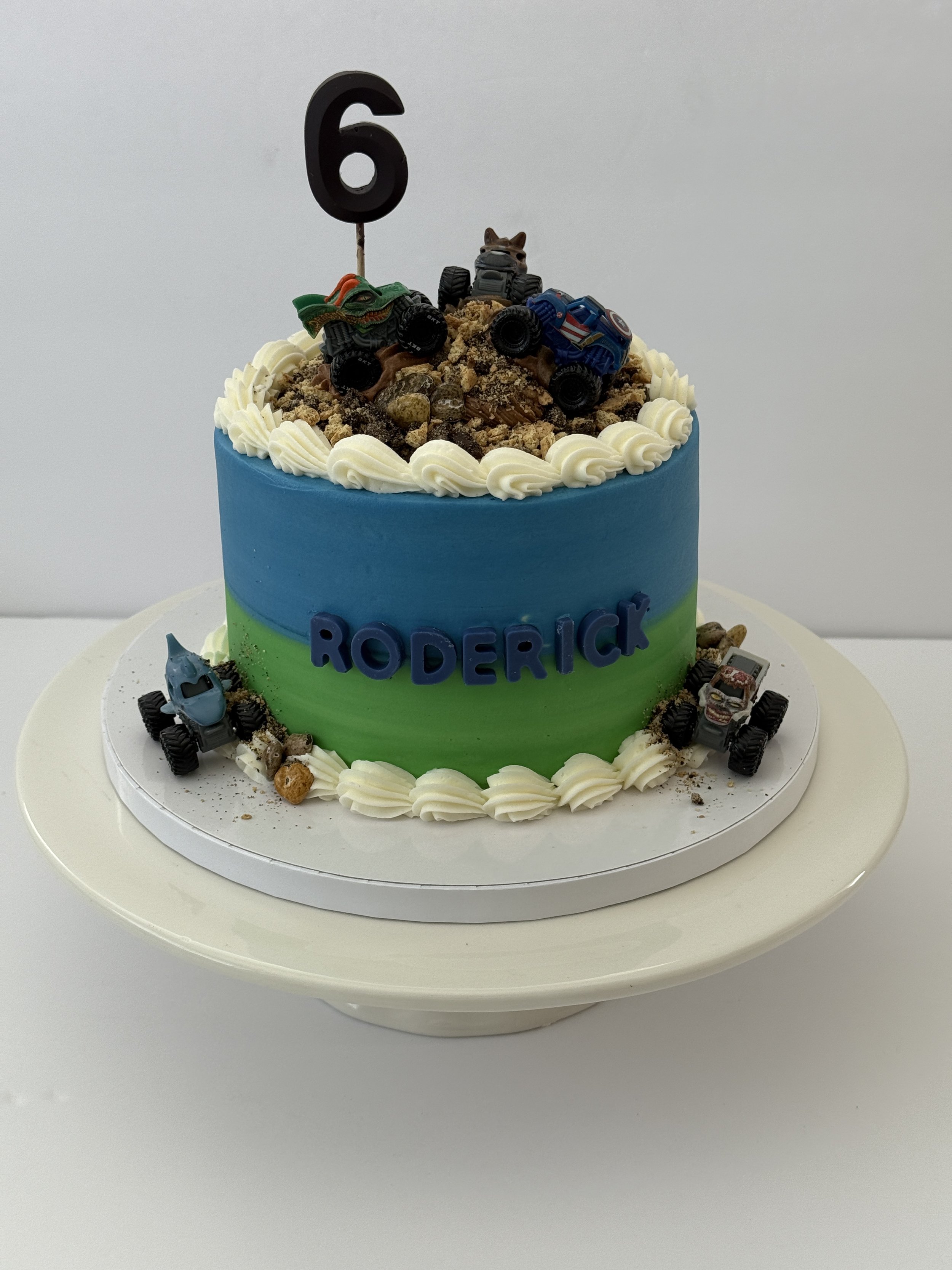 A celebration cake with a blue and green gradient, decorated with white frosting borders, toy monster trucks, and toy cars on top, with a black number 6 candle and the word 'RODERICK' in blue letters.