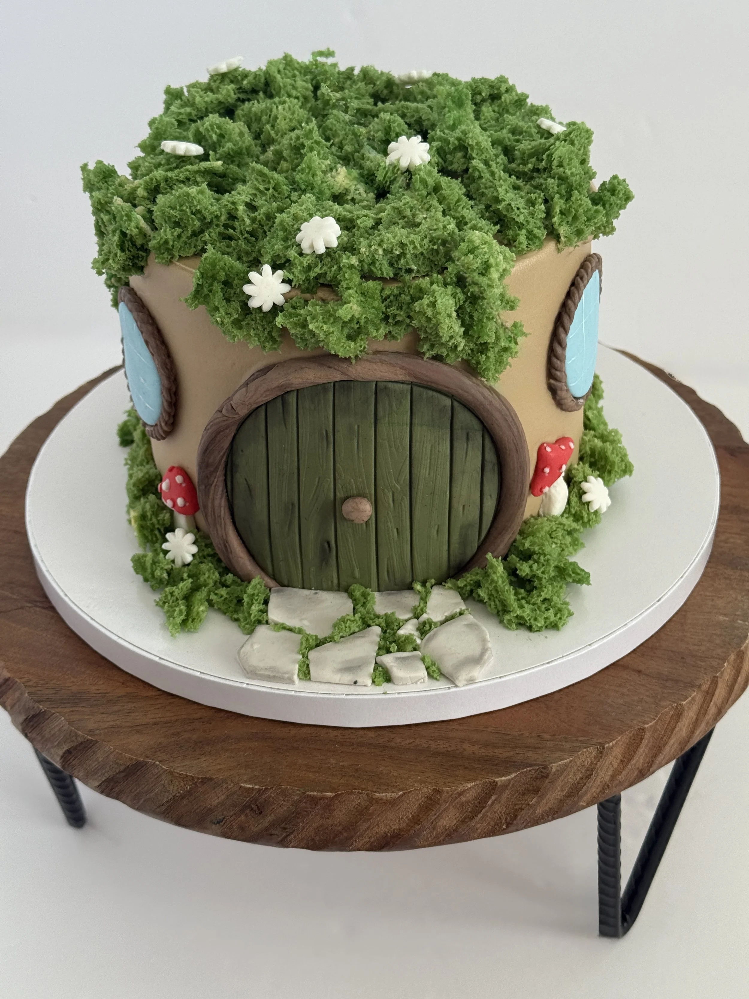 A cake designed to look like a small wooden house with a thatched roof, green grass, and various small decorative flowers, placed on a round white cake board on a wooden table.