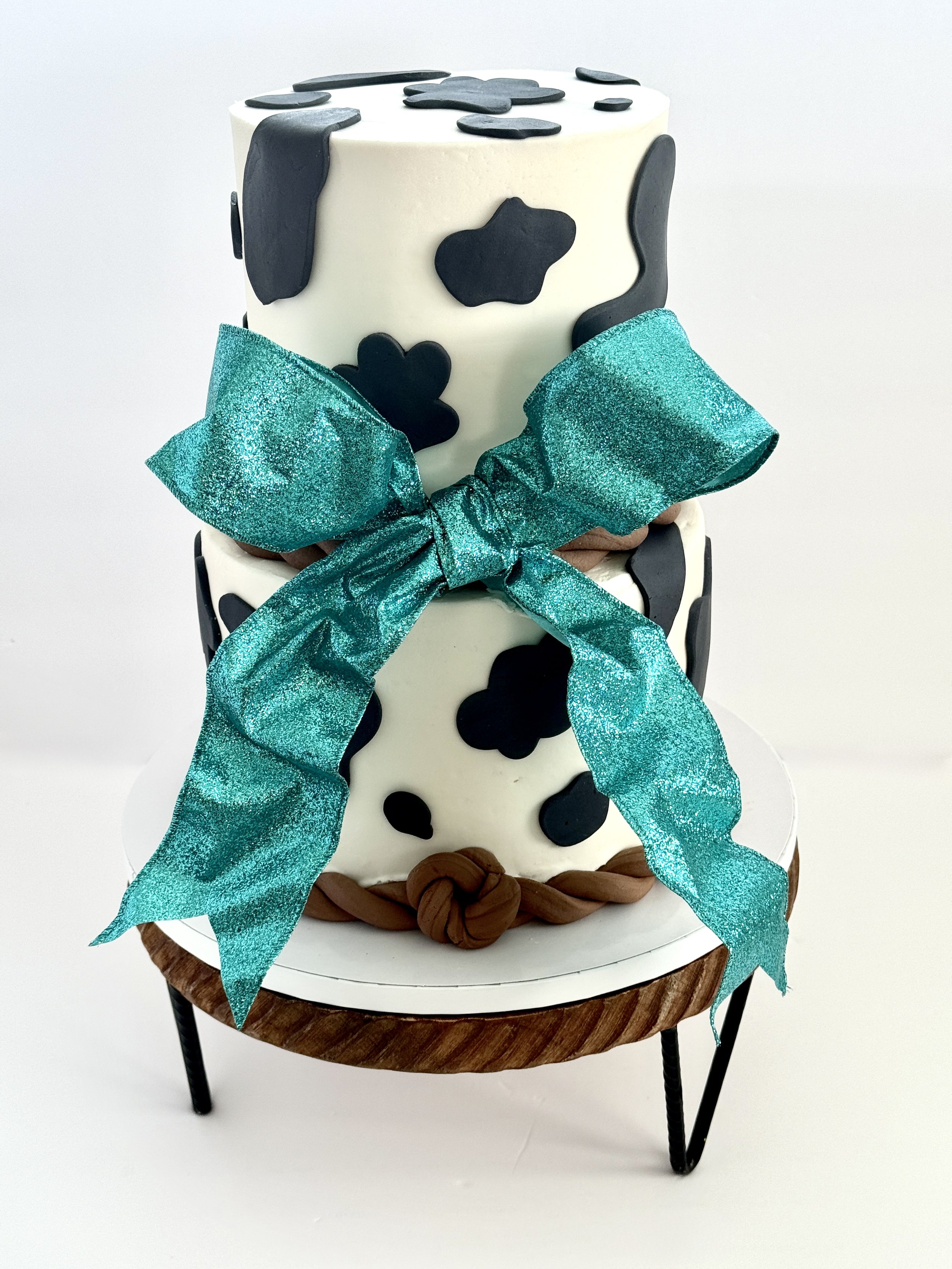 A tiered cake decorated to resemble a cow, with black and white patches, a blue glittery bow, and chocolate accents, placed on a small wooden table.