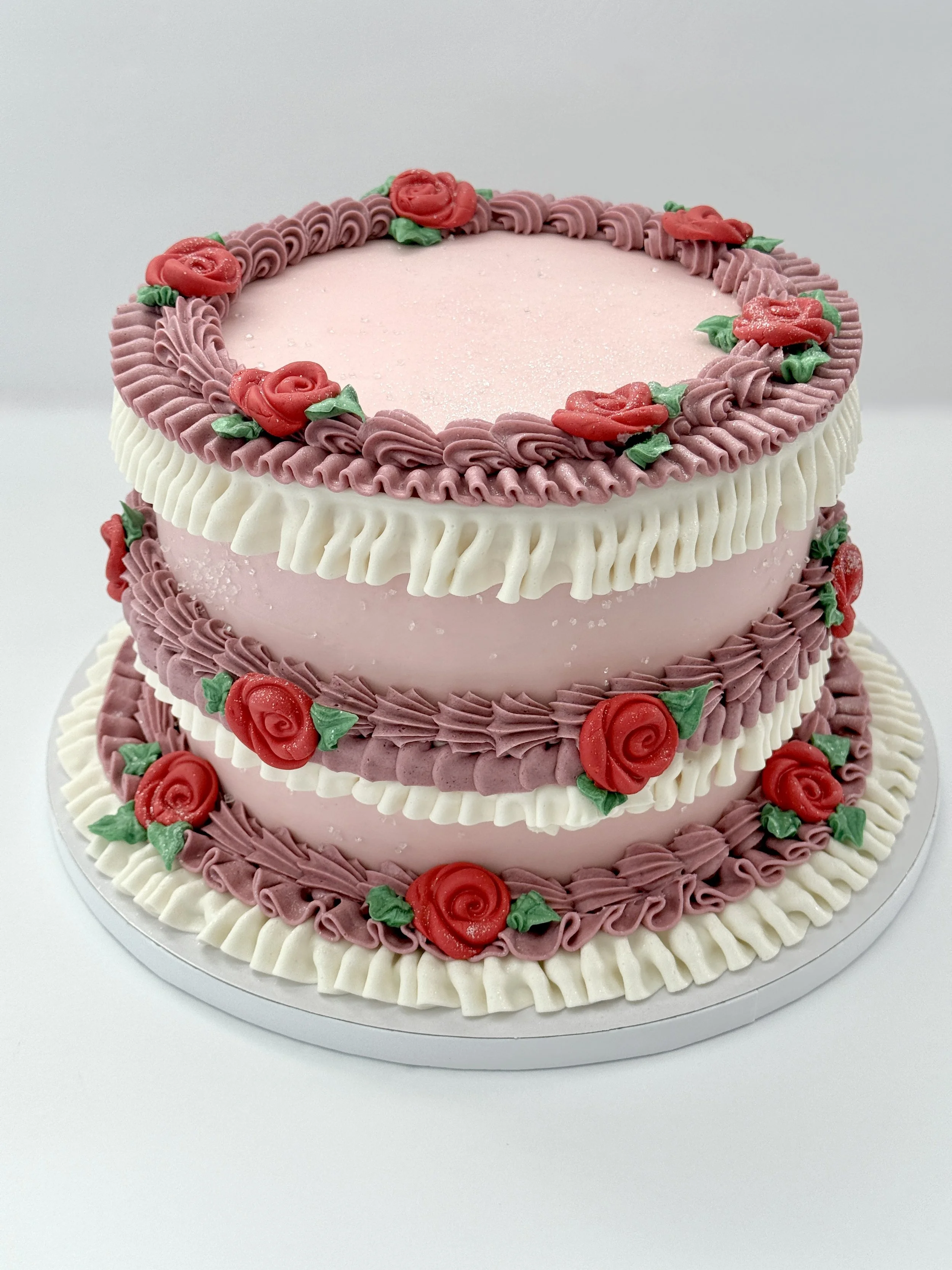 A multi-tiered pink cake decorated with red roses and green leaves made of icing, with cream-colored piping around each layer.