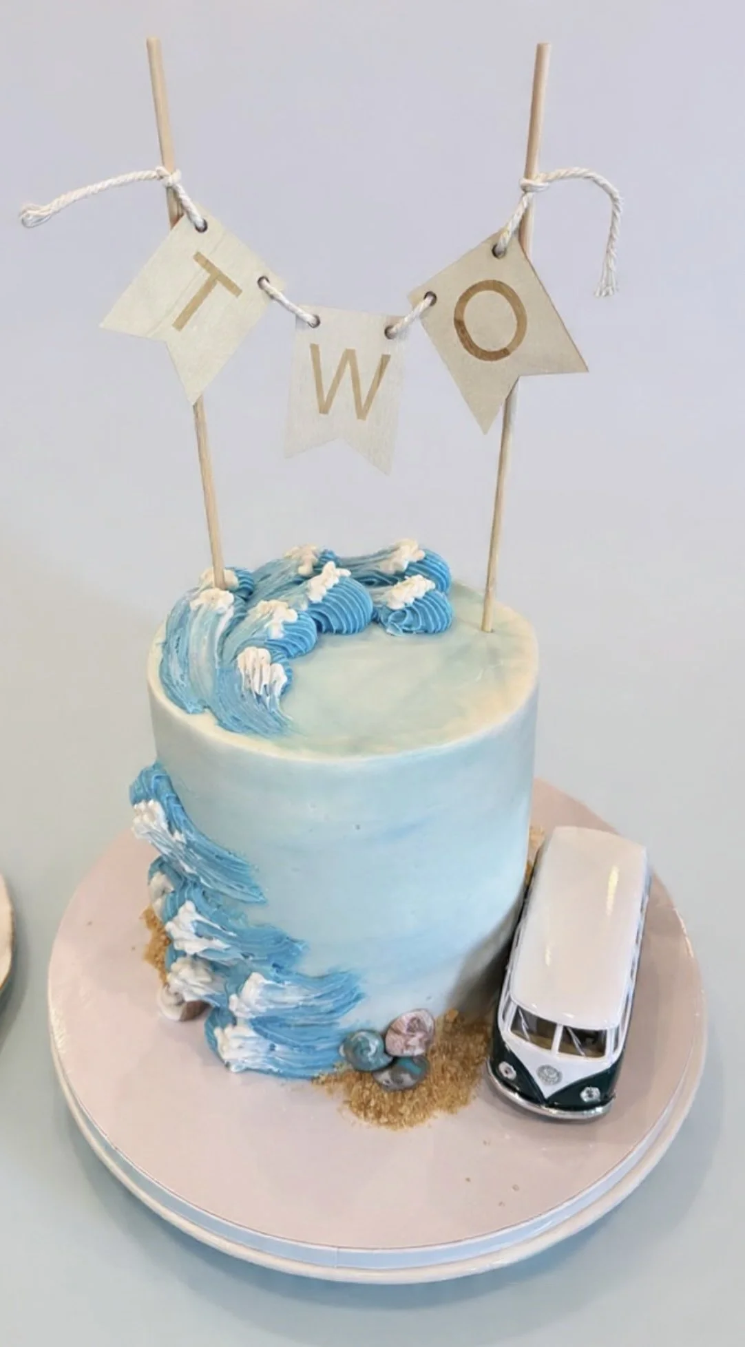 A cake decorated with ocean waves and a miniature black and white van, with a banner that reads "T W O" on top.