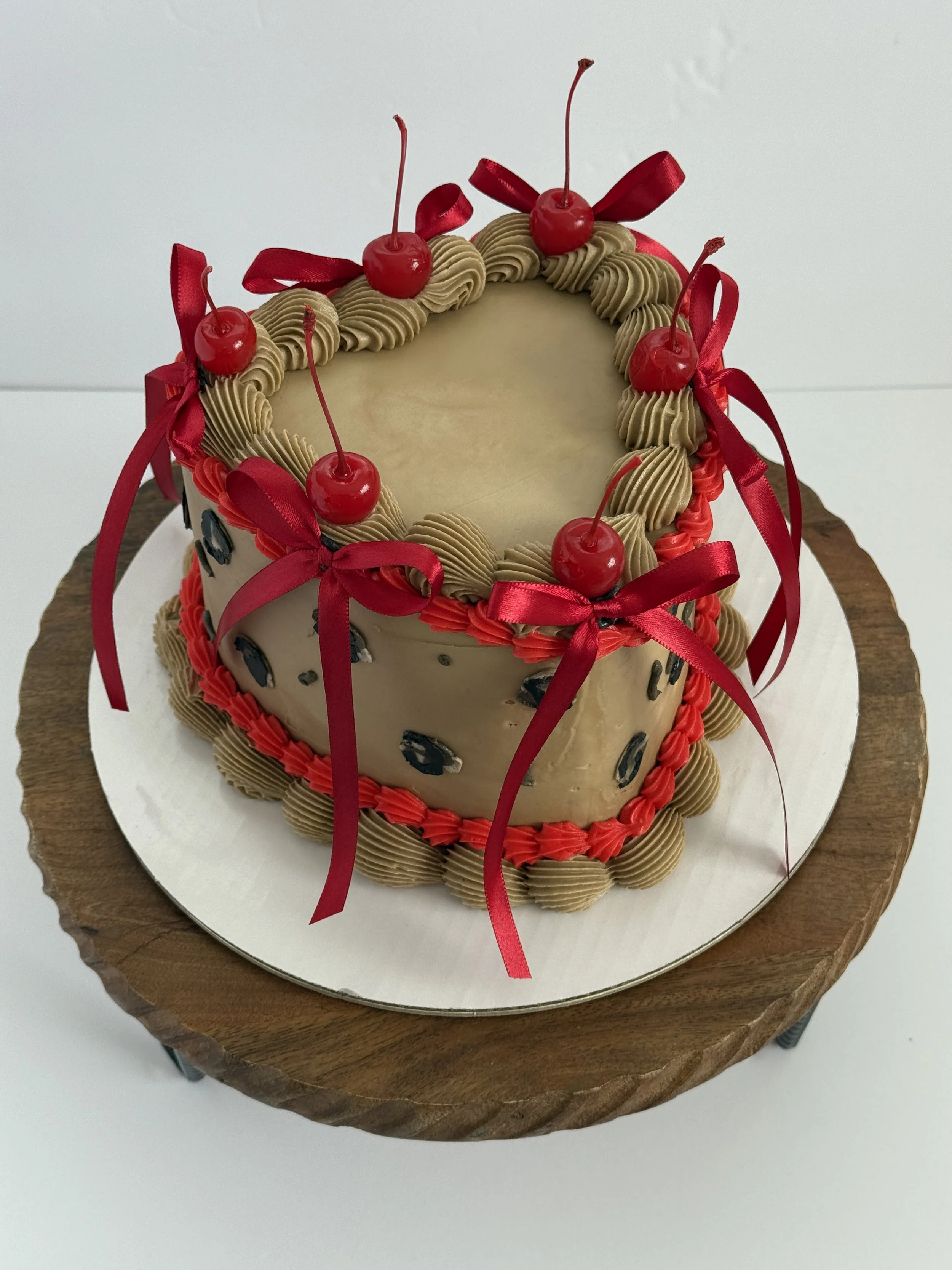 Round cake decorated with cherries and red ribbons on top, and caramel-colored piped borders, set on a wooden cake stand.