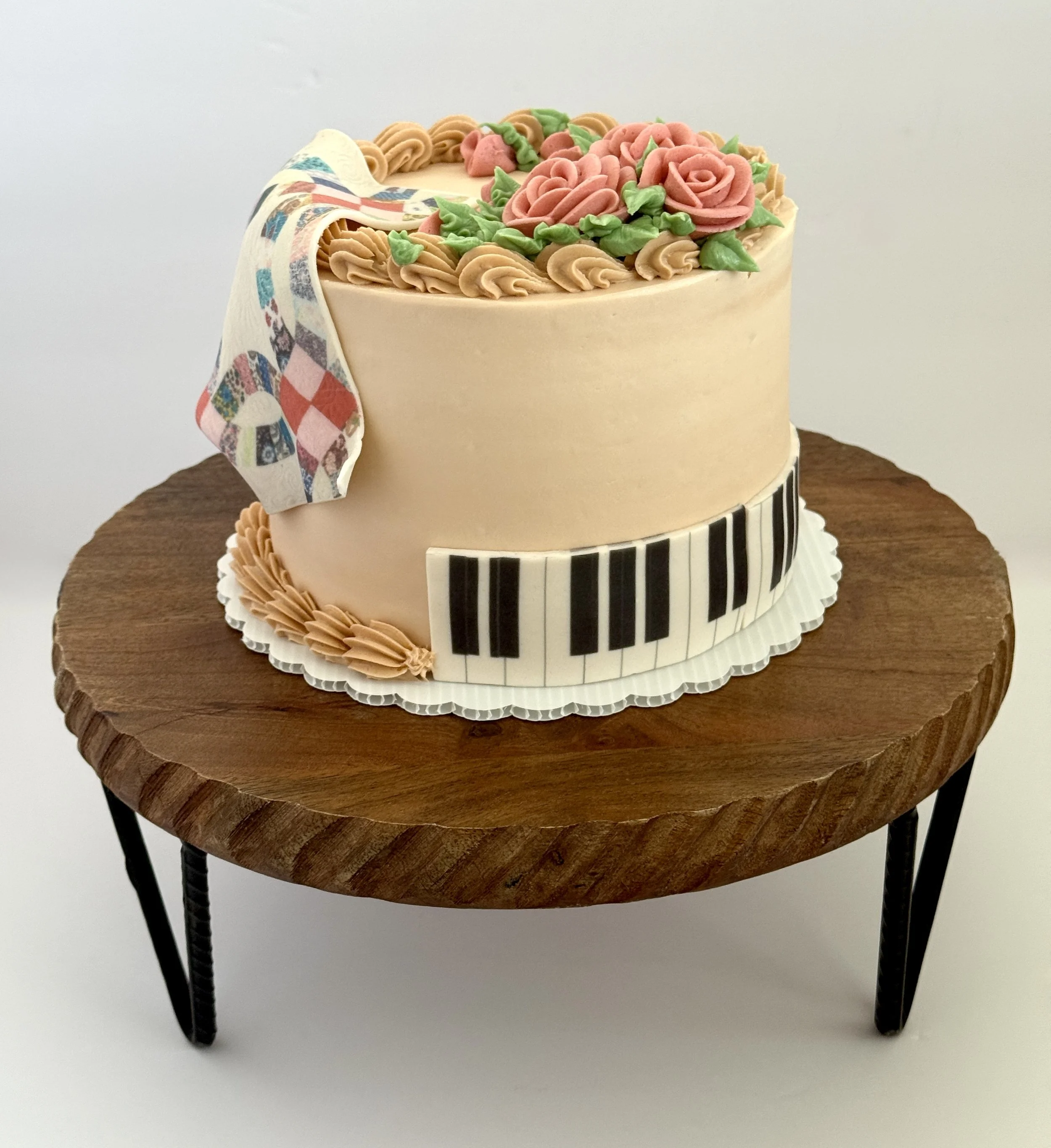 A two-tiered cake on a wooden table with a piano keyboard design around the base, decorated with pink roses, green leaves, and beige frosting accents, with a musical-themed fondant ribbon draped over the side.
