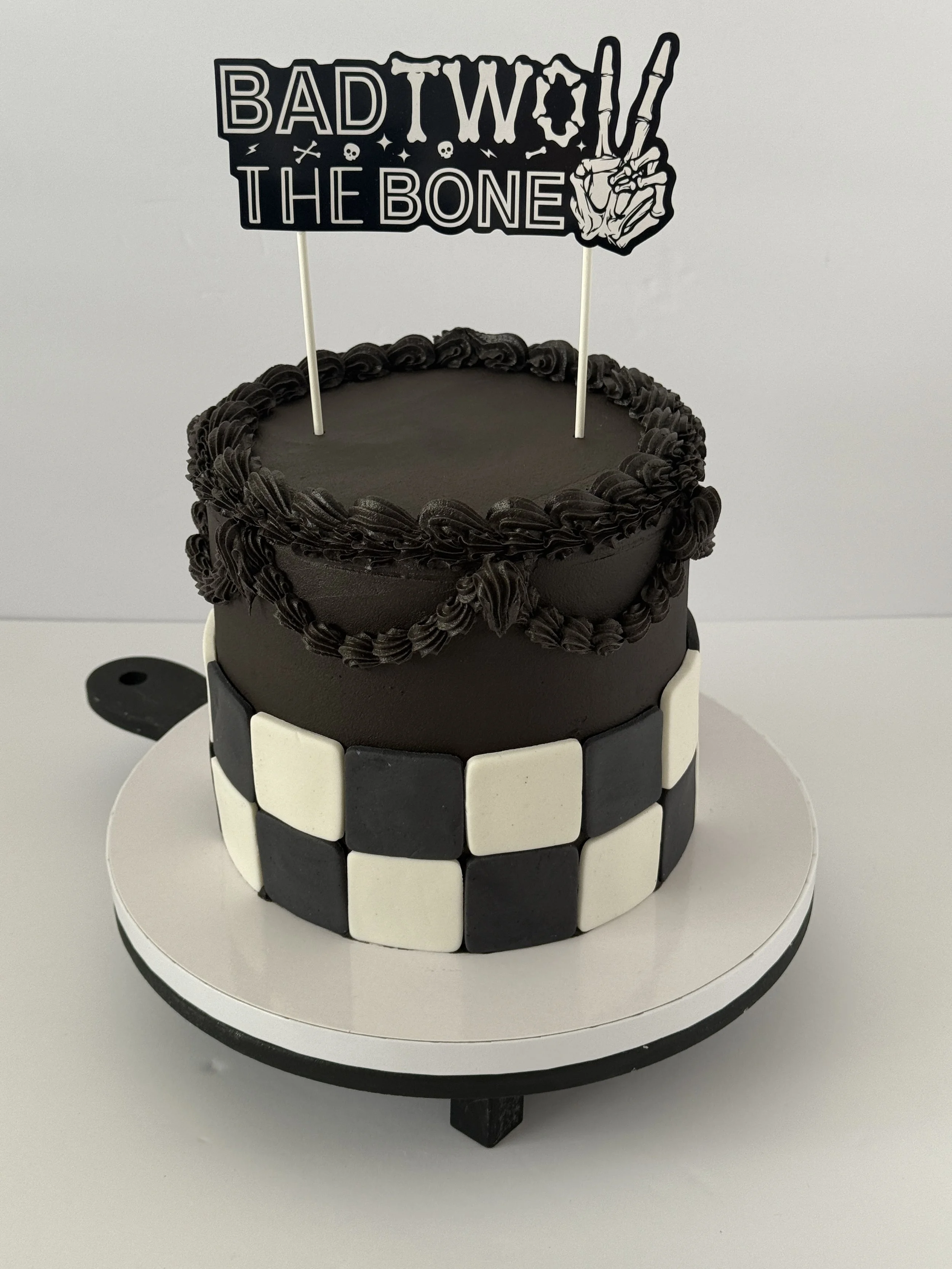 Black cake with a checkered black and white pattern around the sides, sitting on a white cake stand. A black cake topper with the phrase "Bad to the Bone" and a skeletal hand making a peace sign is on top of the cake.