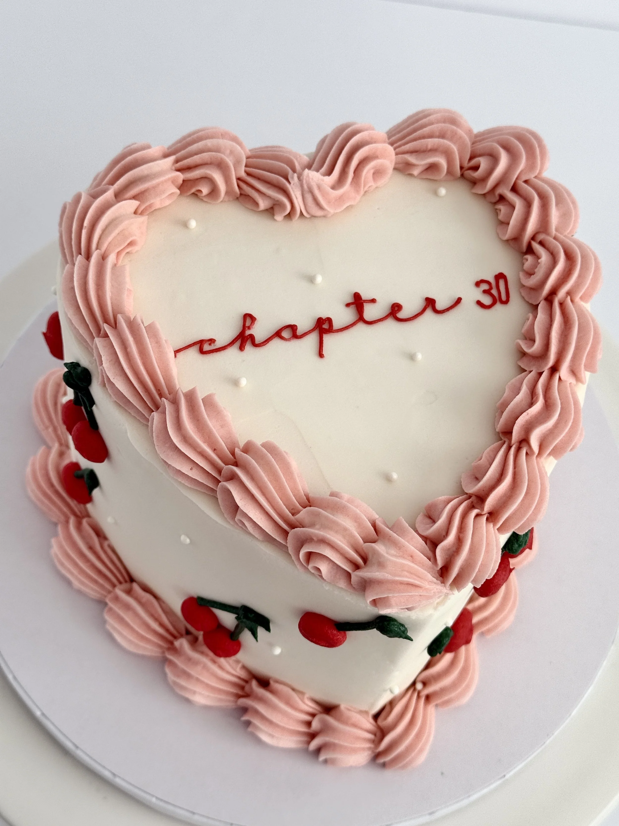 Heart-shaped white birthday cake decorated with pink frosting border, small white edible pearls, and red cherries with green leaves.