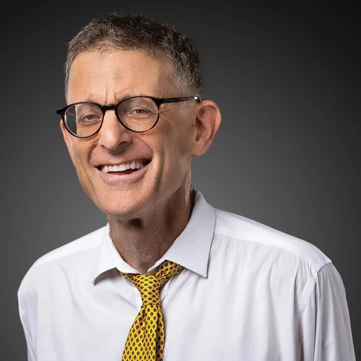 A  man with short gray hair, glasses, wearing a white dress shirt and a yellow tie with black diamond patterns, smiling against a dark background.