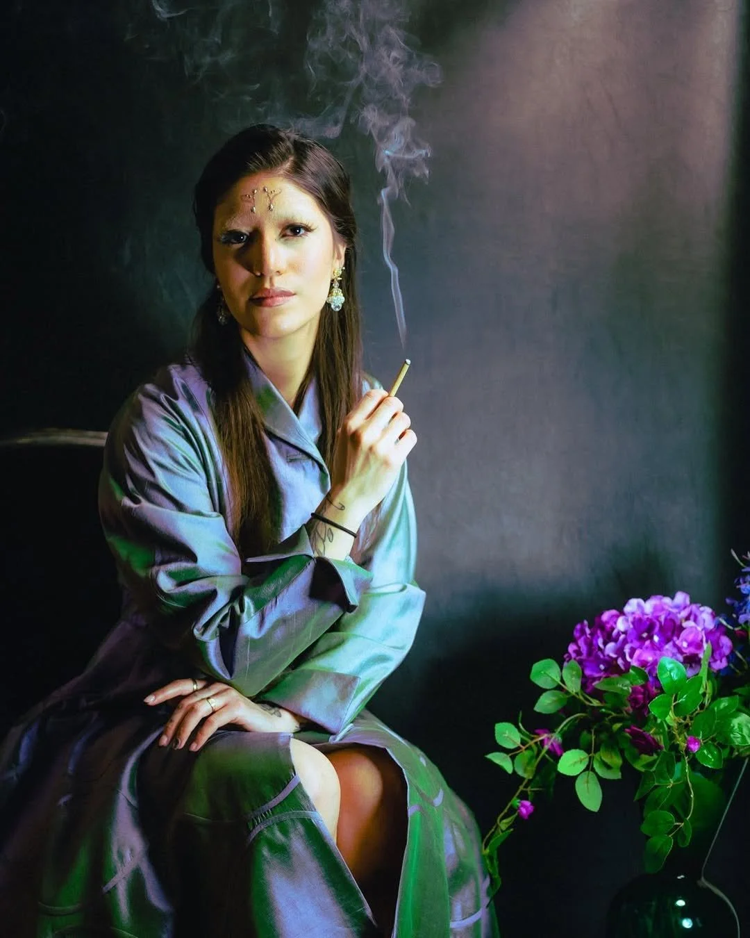 A woman sitting against a dark background, holding a cigarette with smoke rising. She has long dark hair, wears earrings, a jade-colored silk robe, and a black hair tie. There is a purple flower arrangement in a dark vase to her right.