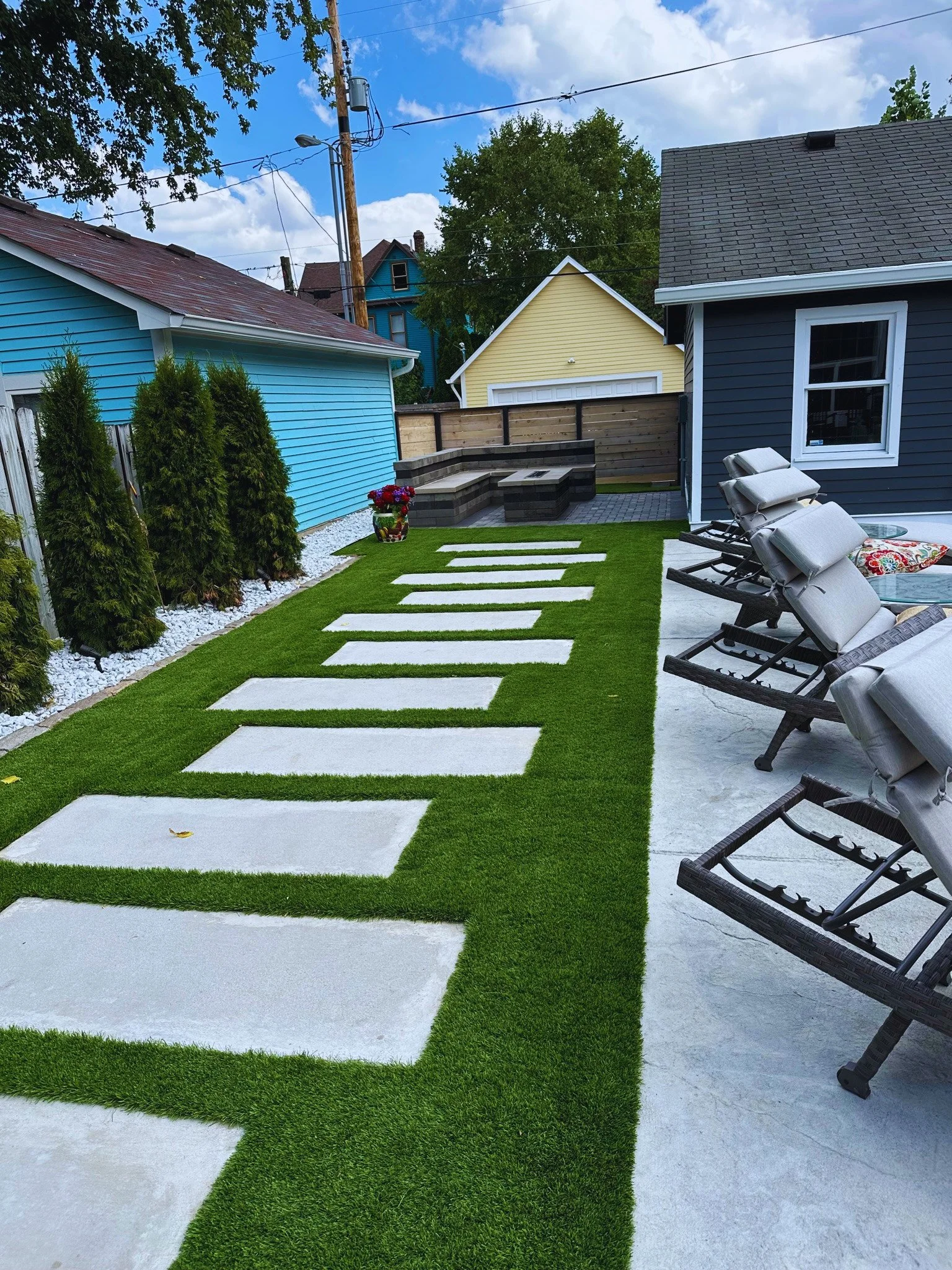 After photo of a modern backyard transformation featuring a custom stone fire pit and seating wall, artificial turf grass with large concrete stepping stones, and luxury lounge chairs.