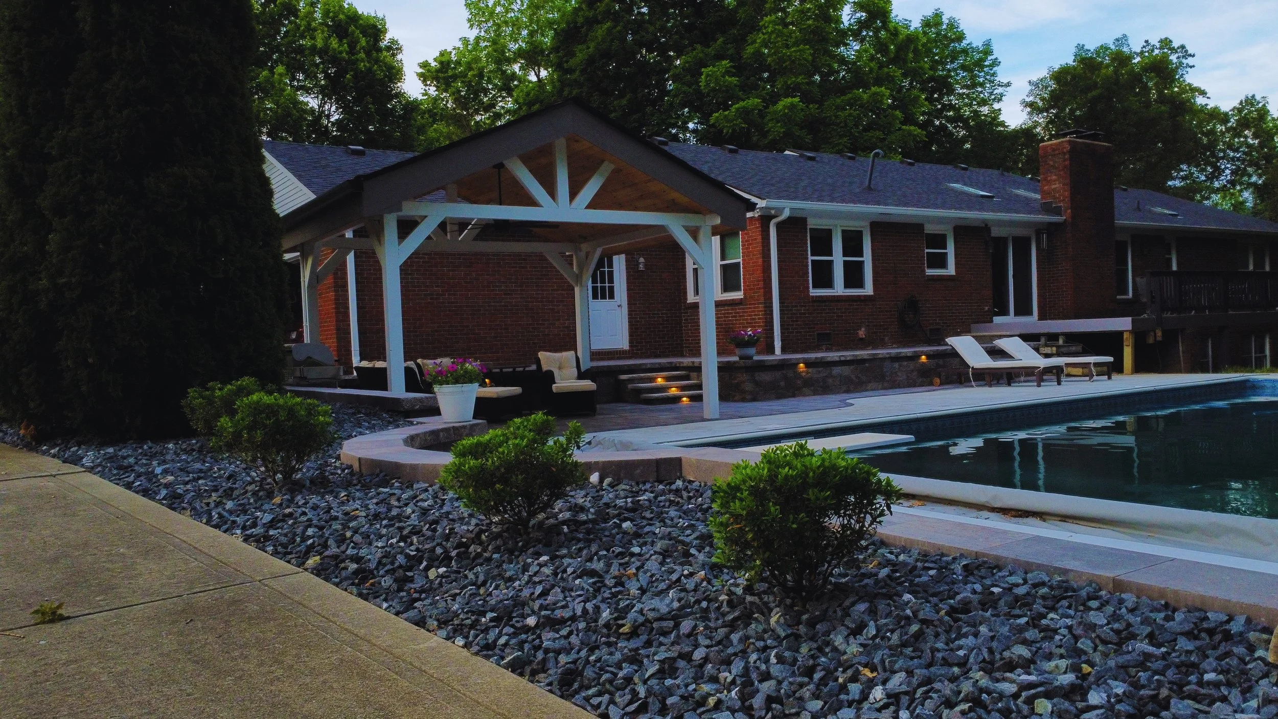 After photo of the completed backyard pool remodel by B&F Landscaping, showcasing a custom white pavilion, premium stone pavers, built-in LED steps lighting, and a modern dark grey river rock landscape border with new shrubs.