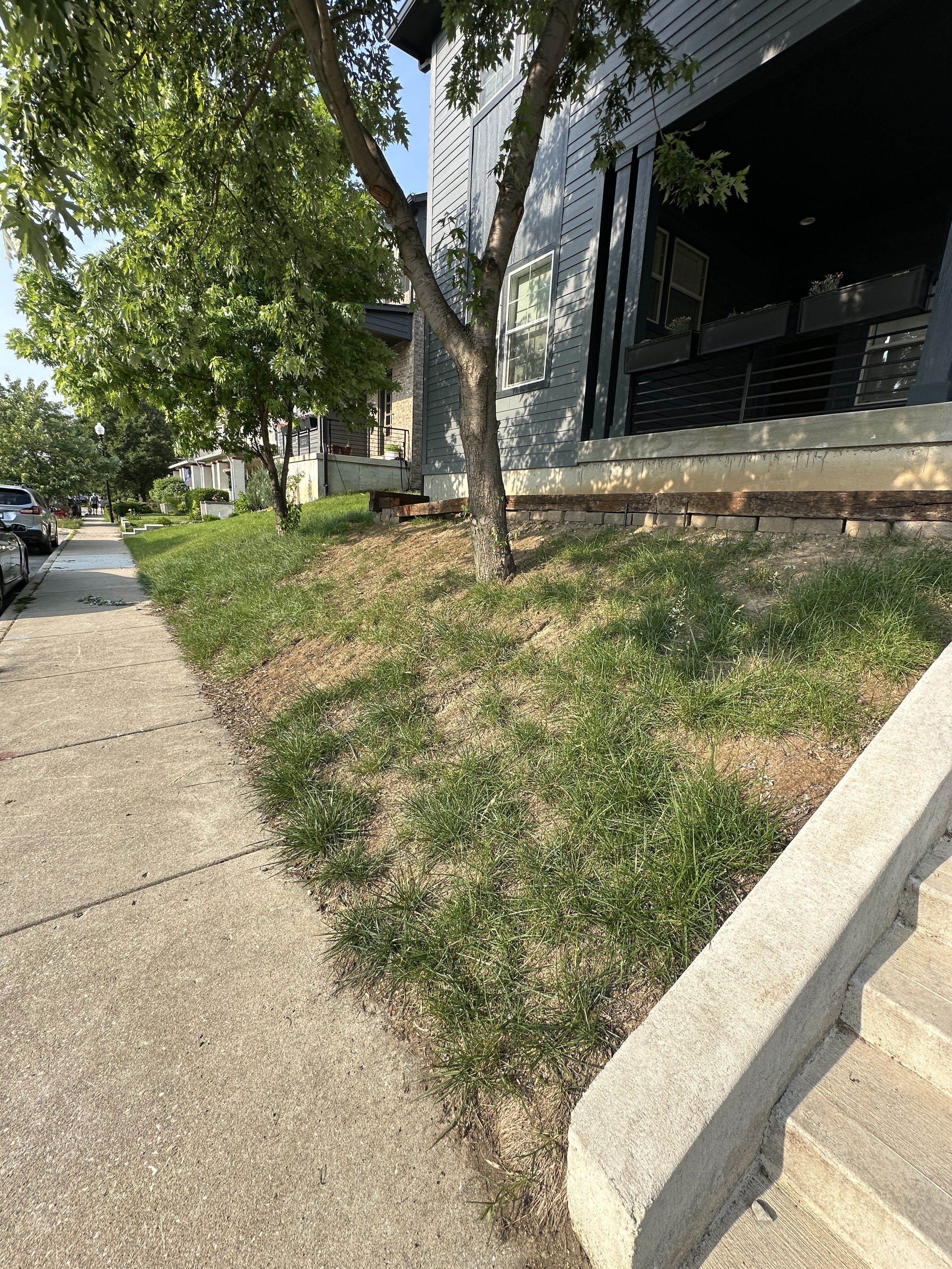 Before photo of a steep sloped front yard with eroding grass and old wooden railroad ties along a city sidewalk, prior to installing a modular block retaining wall.