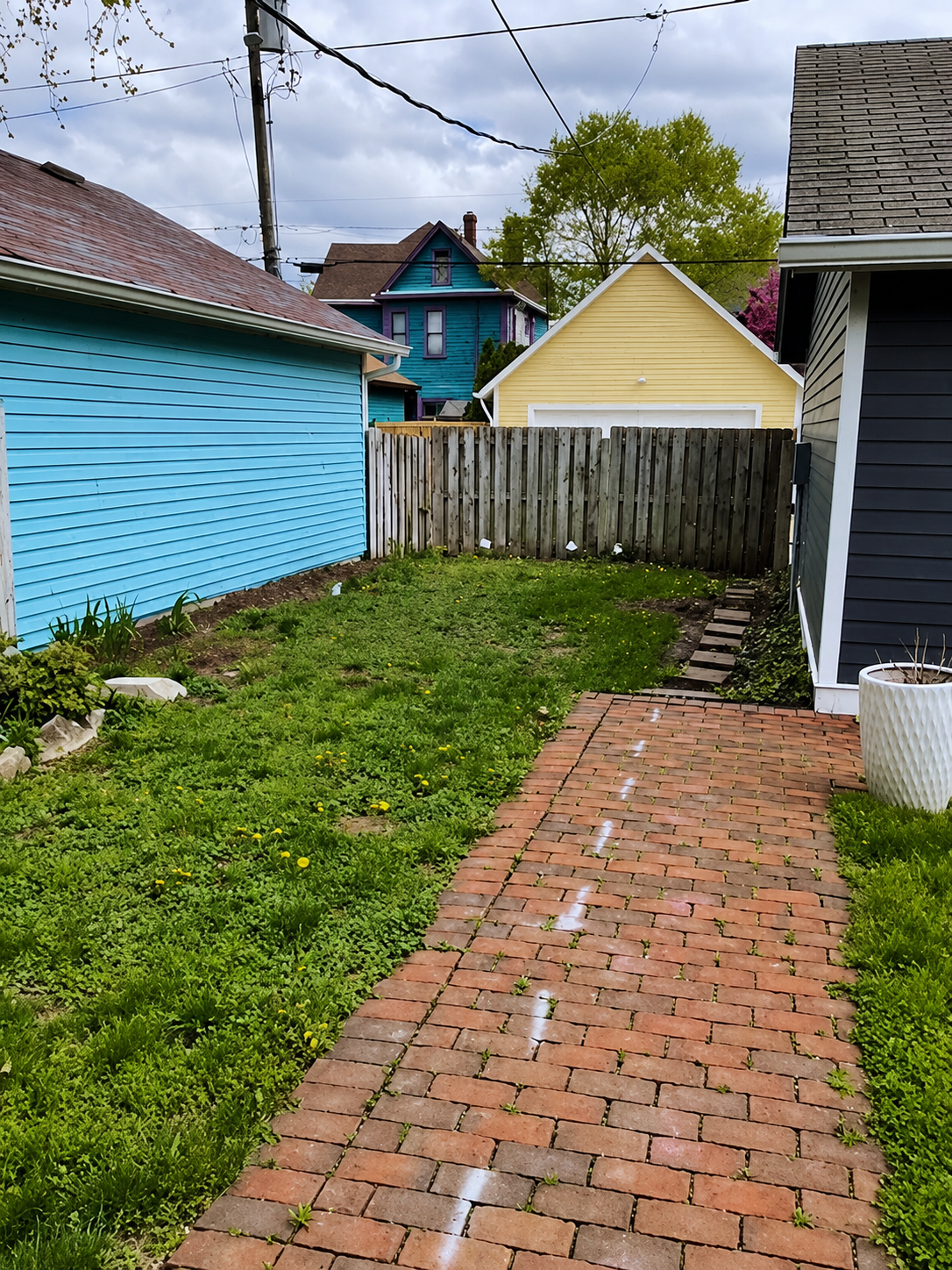 Before photo of a small residential backyard renovation, showing an old brick walkway and grass area before installing a custom paver patio with a fire pit.