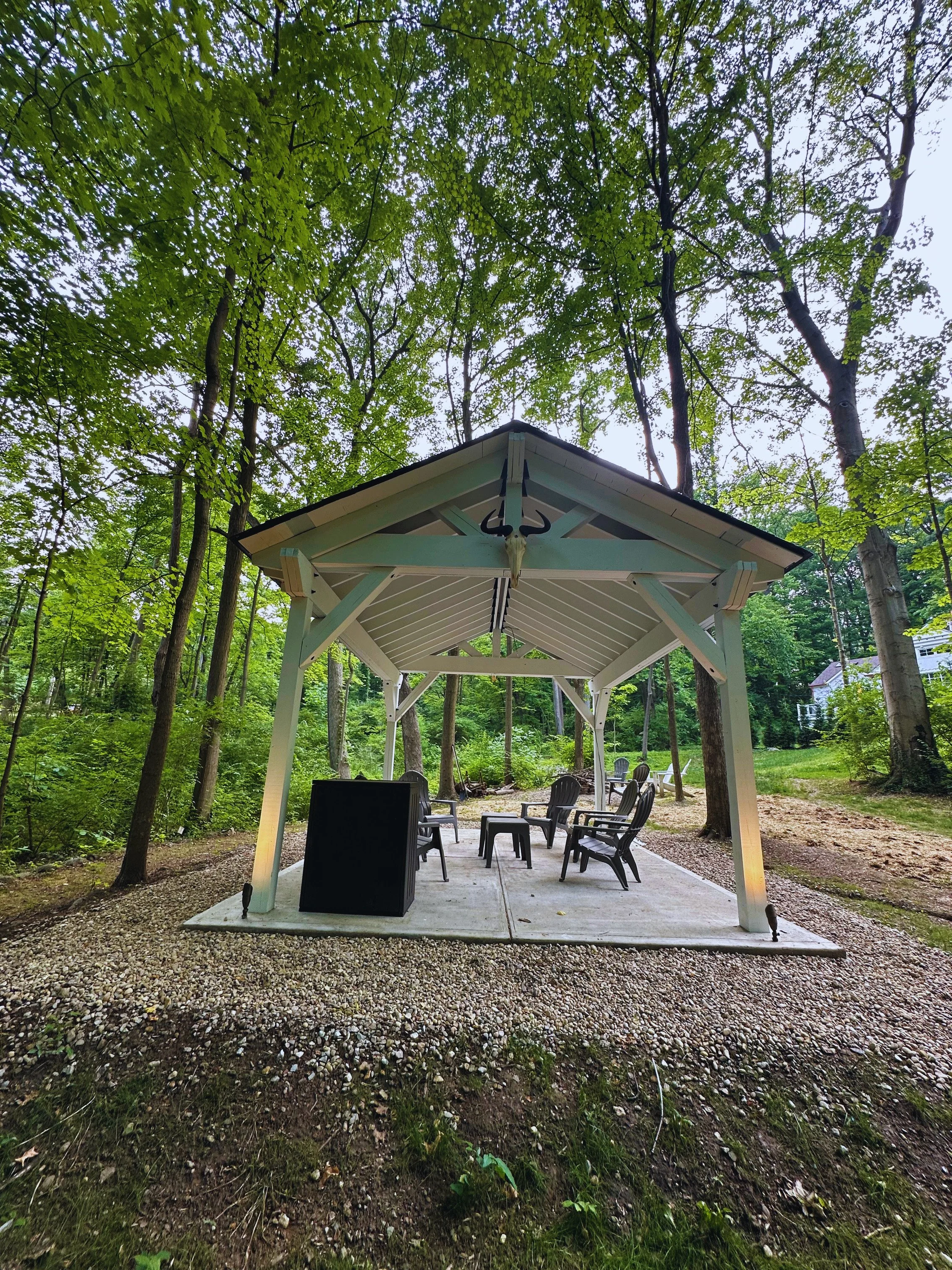 Front view of a custom white detached pavilion with a gabled roof built on a concrete pad by B&F Landscaping, featuring patio chairs in a wooded backyard