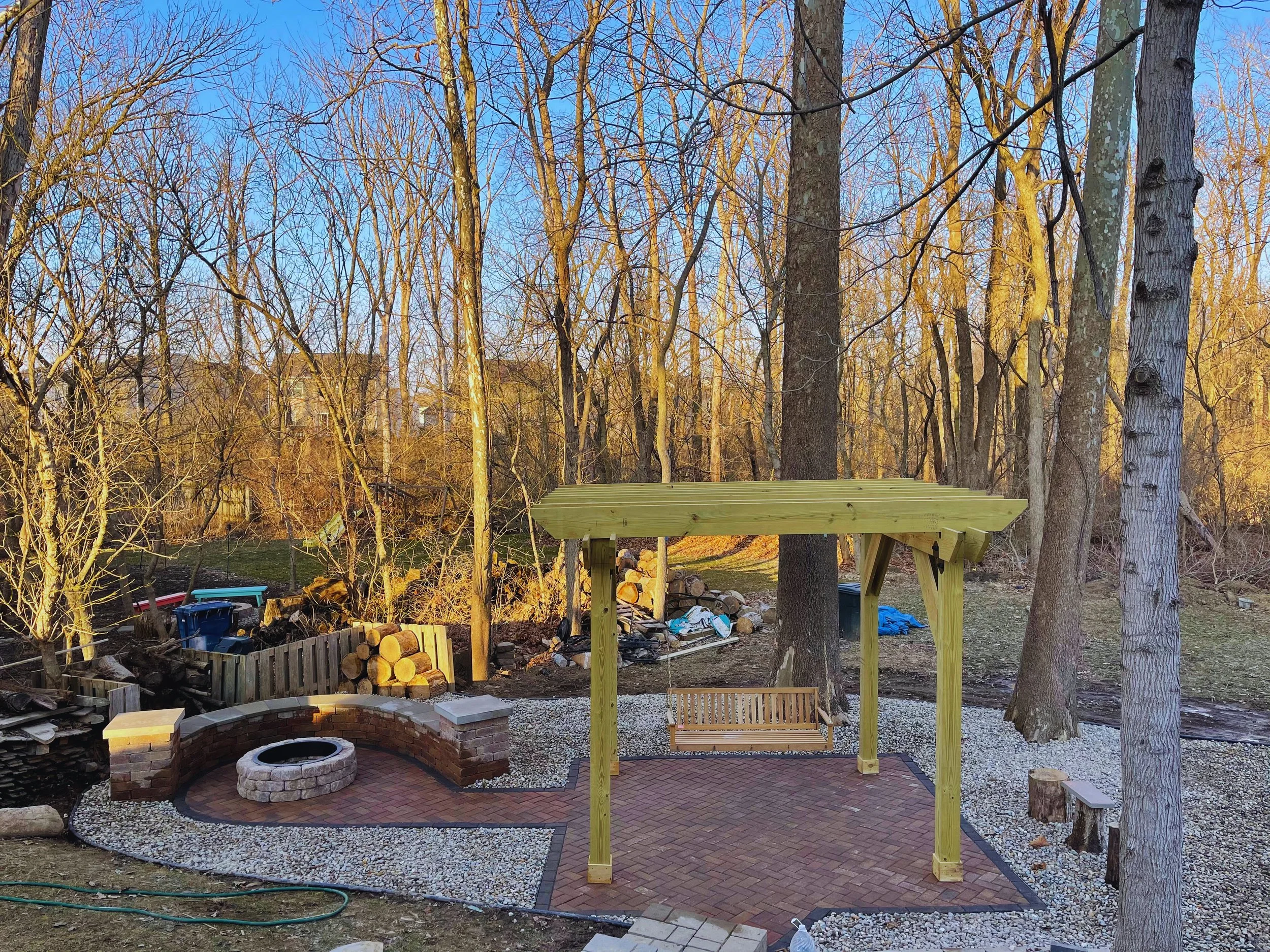 Custom western red cedar pergola with a wooden swing bench, installed by B&F Landscaping over a brick paver patio with a matching stone fire pit.