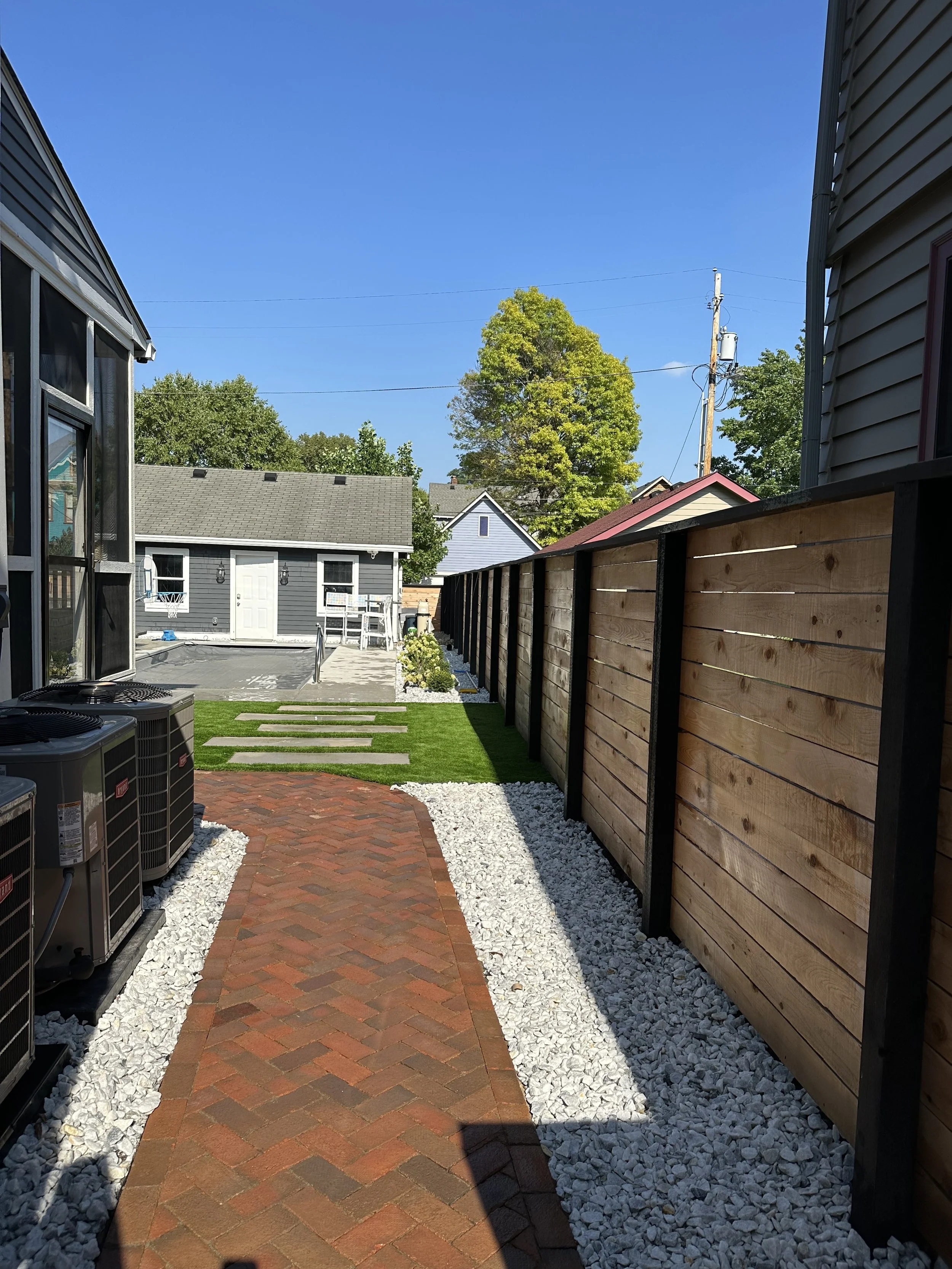 A backyard walkway with brick pavers, white gravel borders, leading to a grassy area with stepping stones. On the left are air conditioning units, and on the right is a wooden fence. In the background, there is a small gray building, trees, and houses under a clear blue sky.