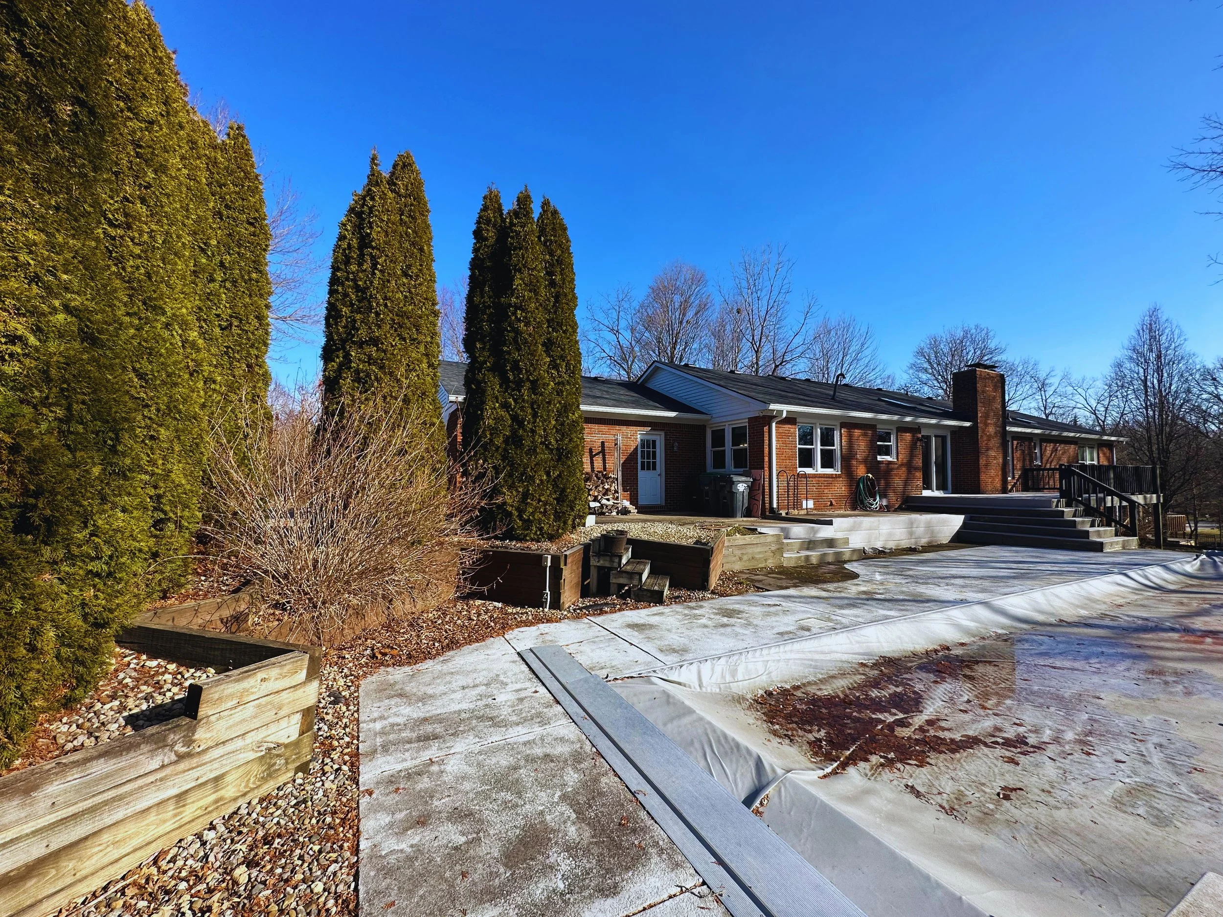 Before photo of a backyard pool landscaping project, featuring old wooden retaining walls, an outdated concrete pool deck, and overgrown bushes before B&F Landscaping's remodel.