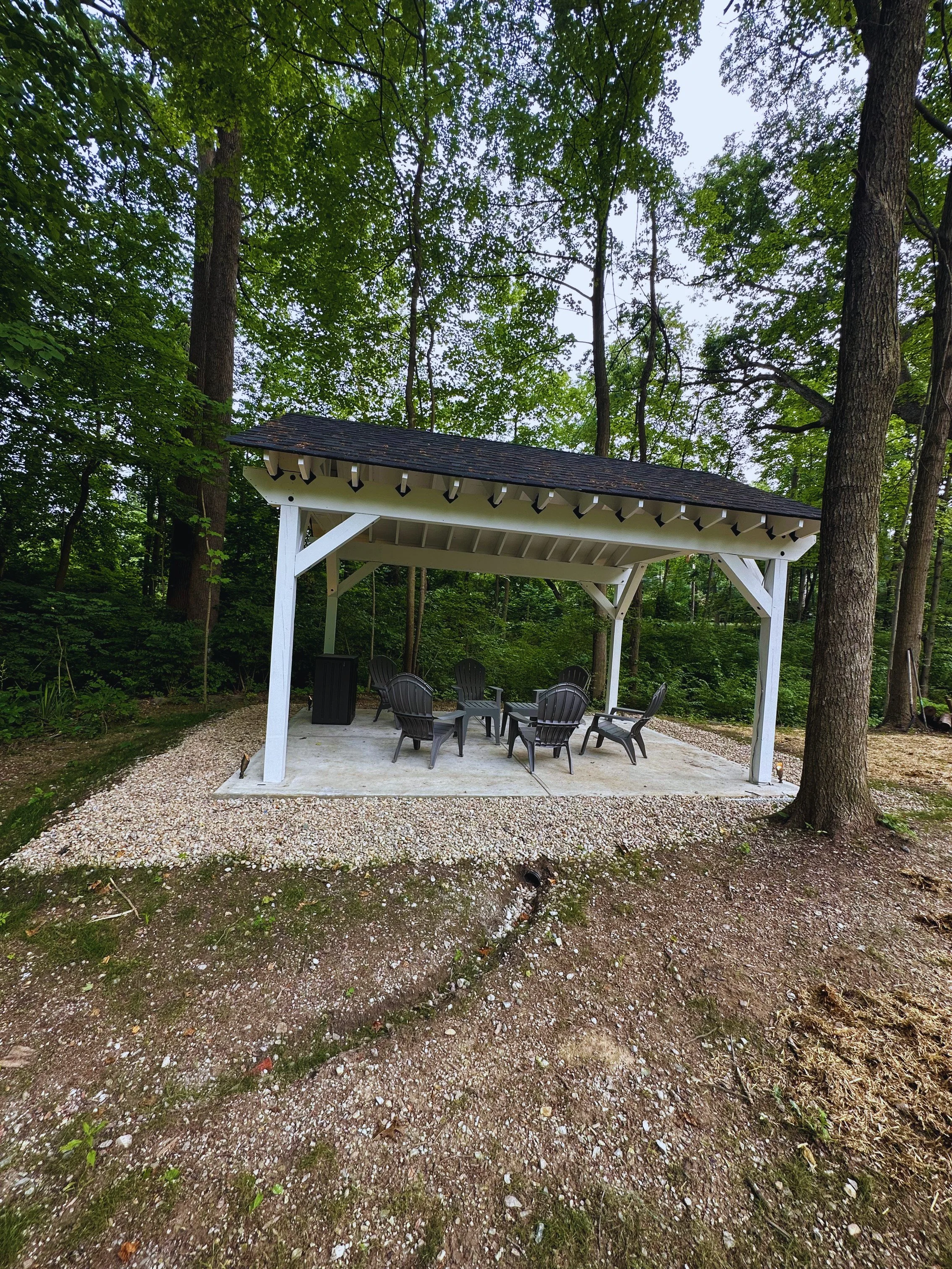 Side angle of a white solid-roof pavilion structure surrounded by a river rock gravel border and mature trees, showcasing a secluded outdoor seating area.