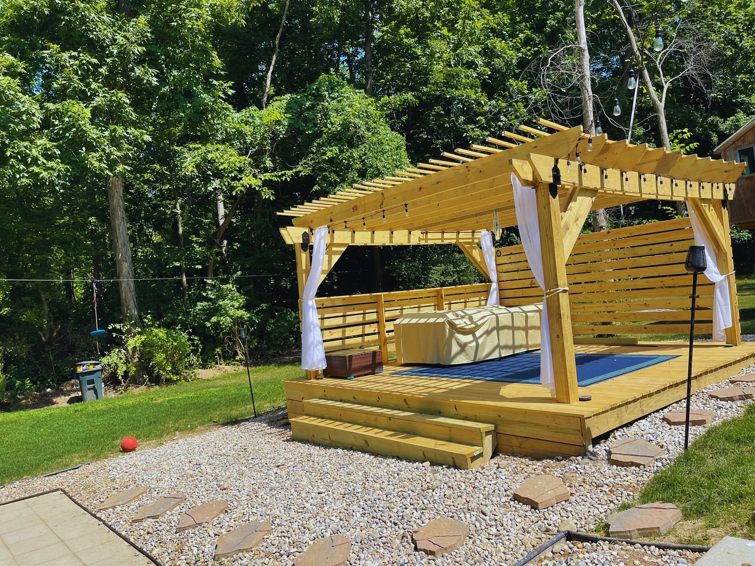 Custom wooden freestanding pergola with a privacy slat wall and white curtains, built over a raised wood deck by B&F Landscaping.