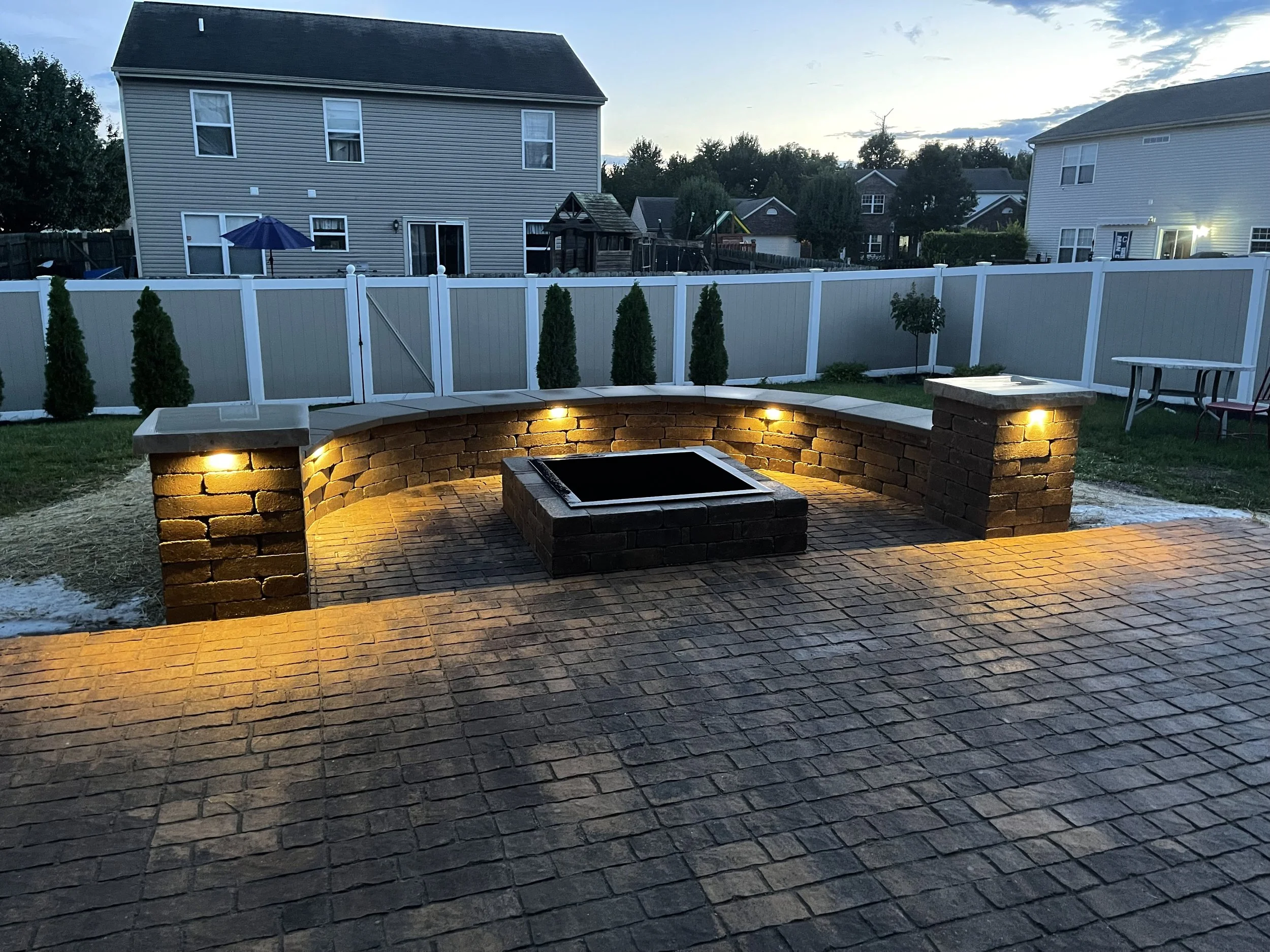 Custom paver patio installation featuring a built-in square stone fire pit, curved seating wall with integrated LED lighting, and pillars in Indianapolis.