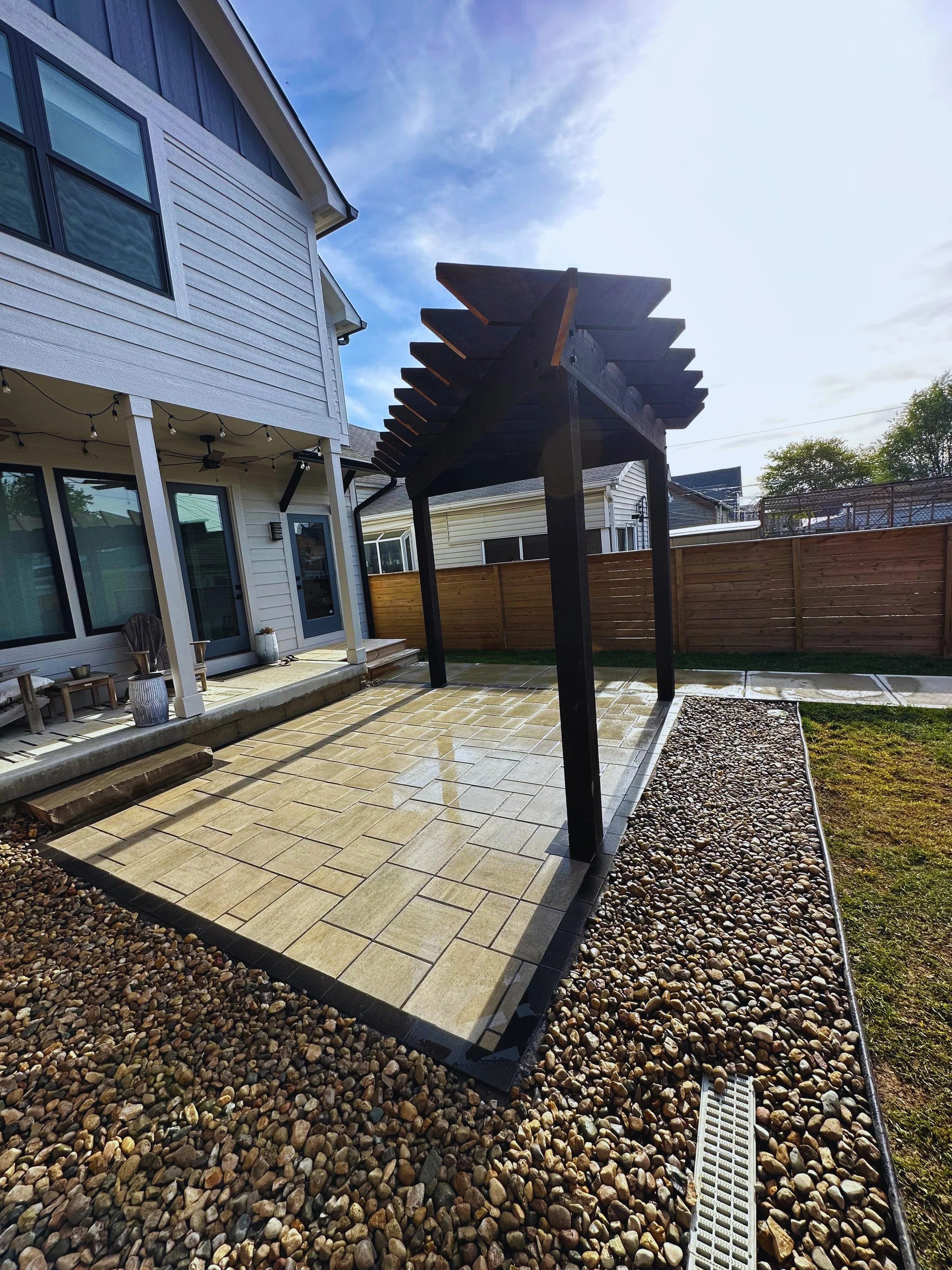 Close-up view of a dark-stained freestanding wooden pergola built over a modern tan paver patio with a wide river rock drainage border by B&F Landscaping.