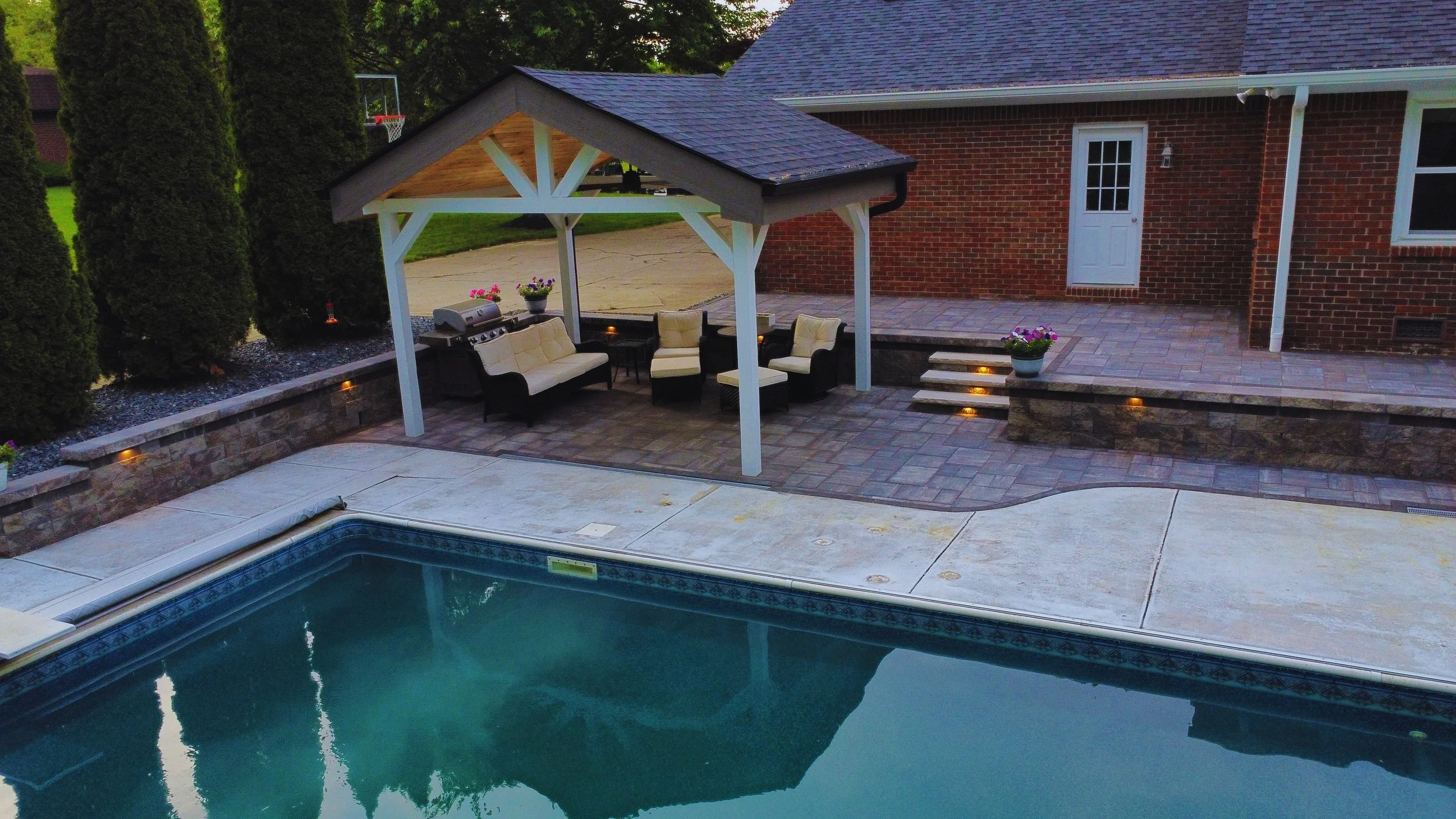 Traditional backyard pavilion with a solid gabled roof and white pillars, built over a custom paver patio next to a swimming pool by B&F Landscaping.