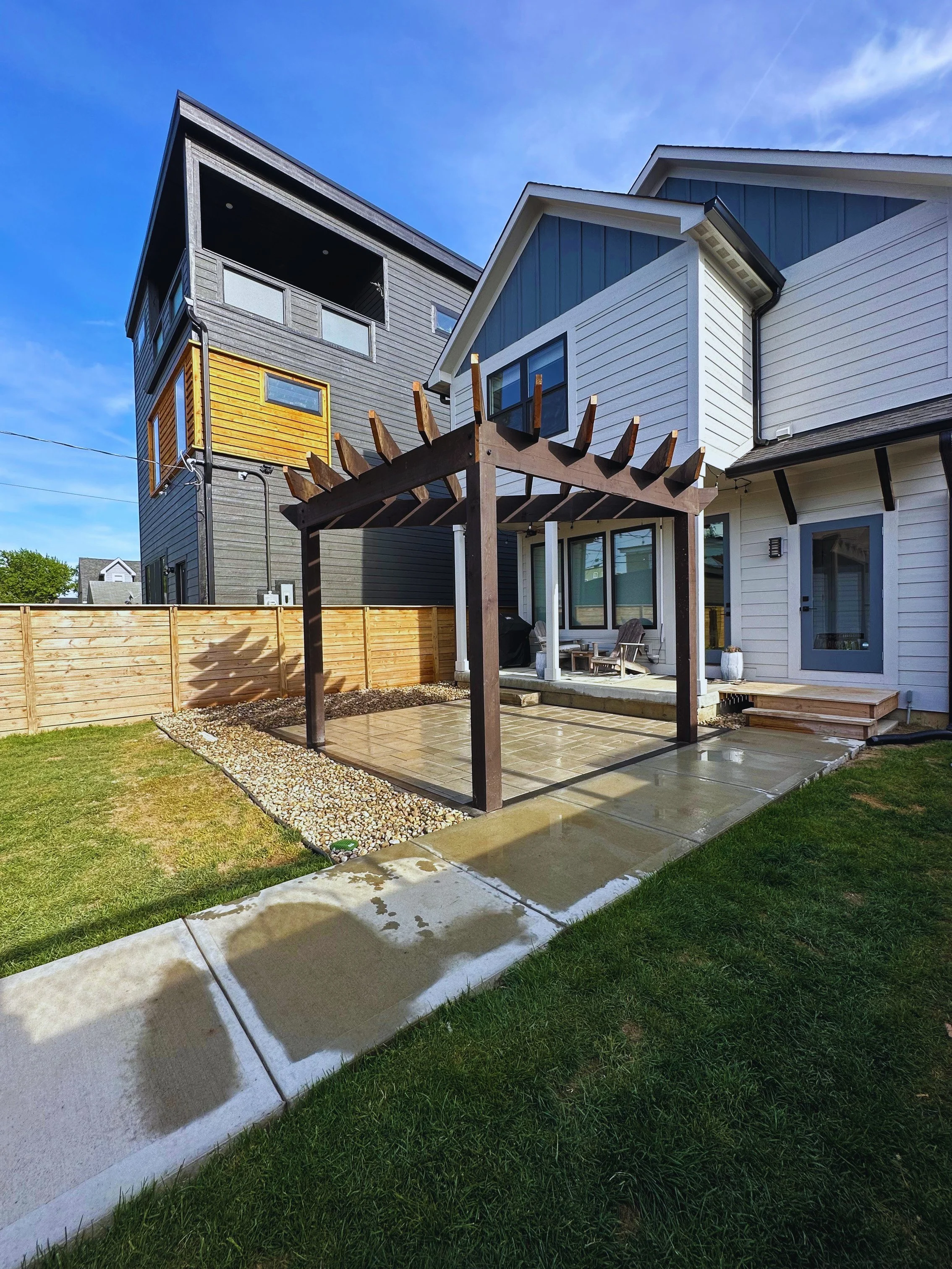 Full backyard landscape view showing a dark wood pergola on a paver patio extension, a concrete walkway, and a wooden privacy fence next to a modern home.