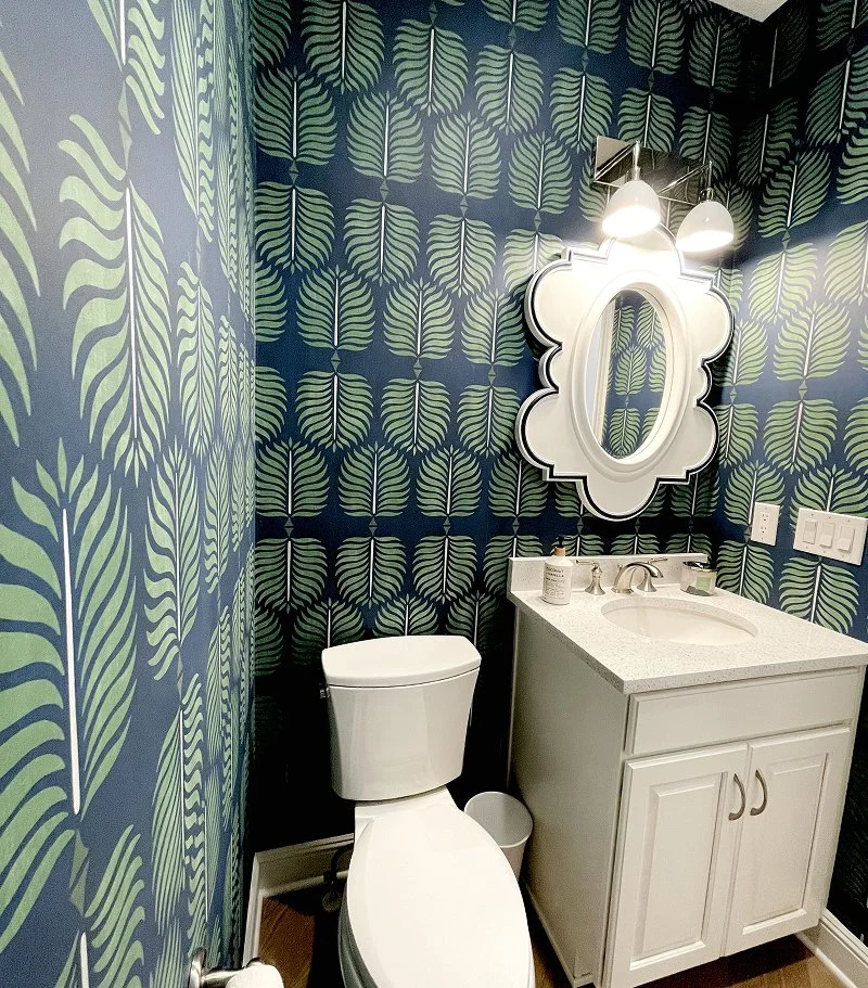 Stylish half bath designed by Interior Designers in Denver, CO, featuring dark teal leaf wallpaper, white fixtures, and round mirror.