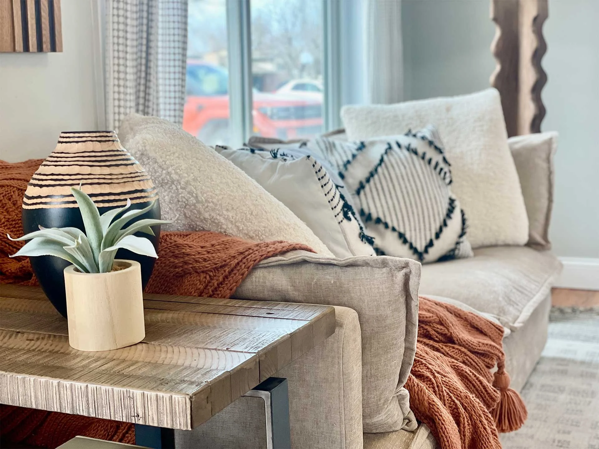 Cozy living room corner with plush beige sofa, pillows, orange throw blanket, wooden side table, potted plant, and vase, showcasing residential home staging services.