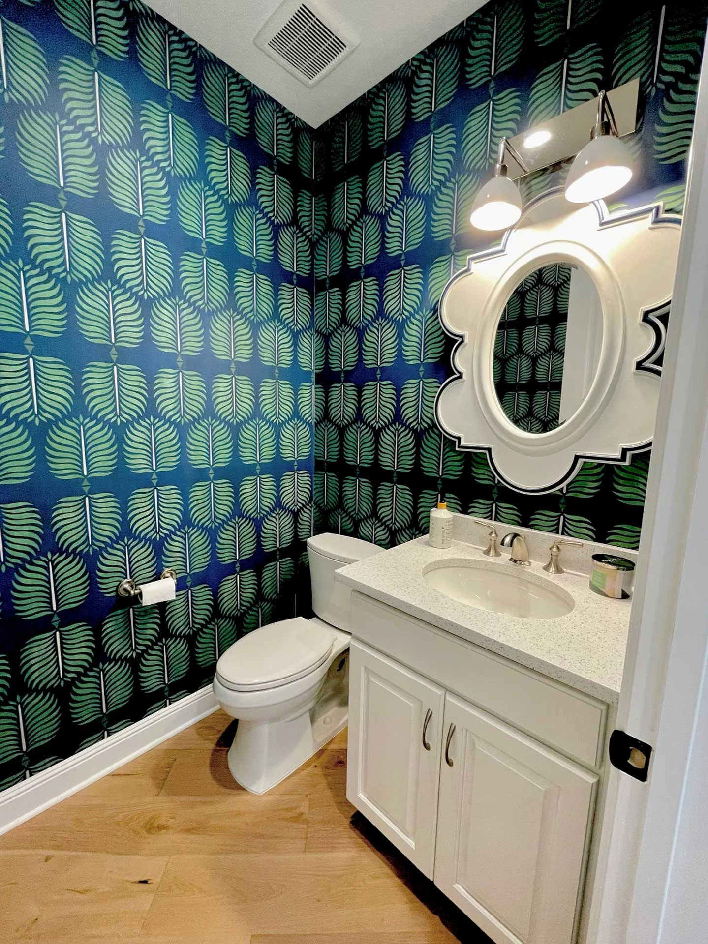 Powder room with bold blue and green leaf-pattern wallpaper, white vanity with speckled countertop, oval mirror, and modern double sconce lighting.