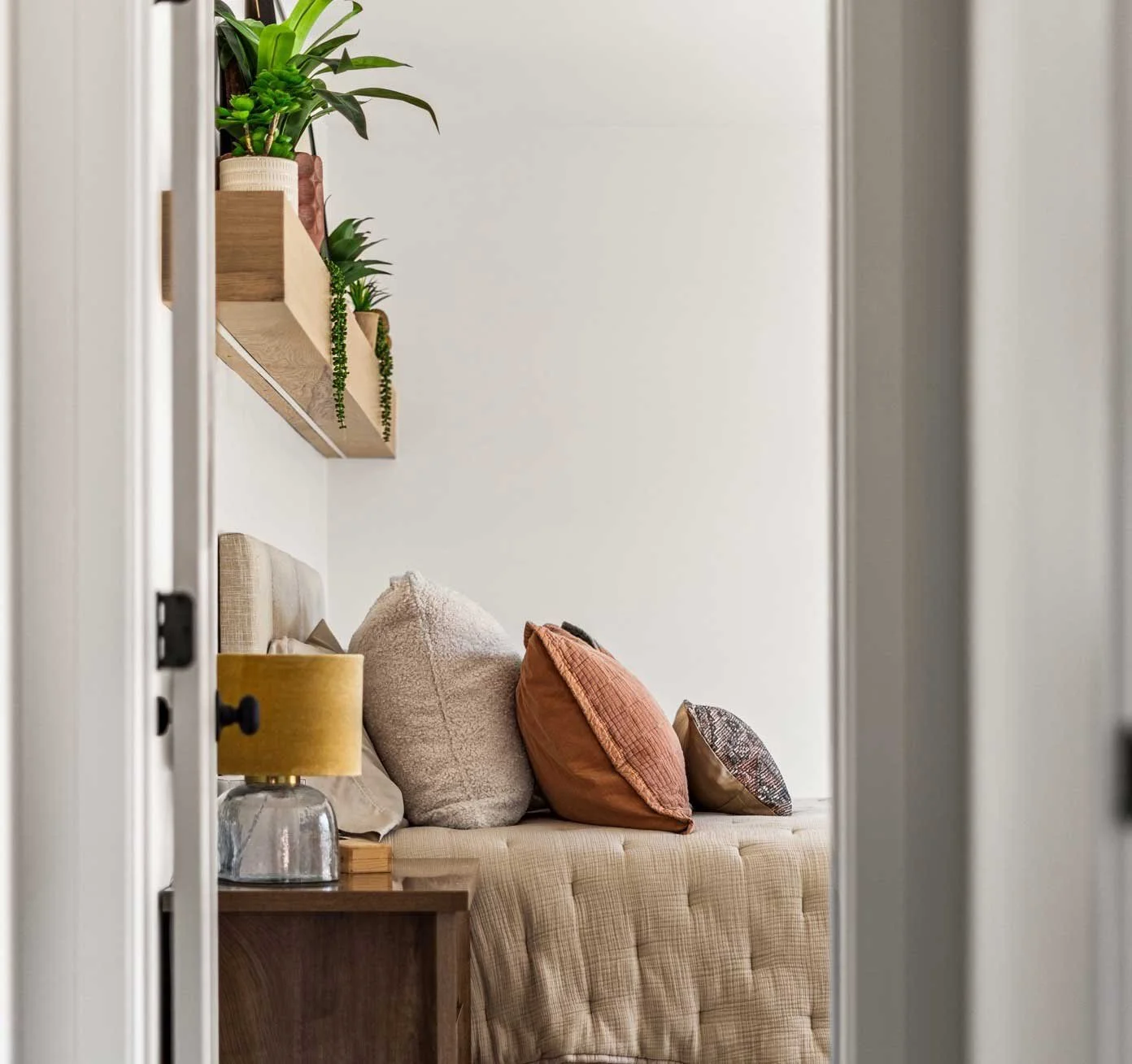 Elegant bedroom setup: quilted bed, layered cushions, plants atop wood shelves, yellow lamp, styled by interior decorators in Denver CO.