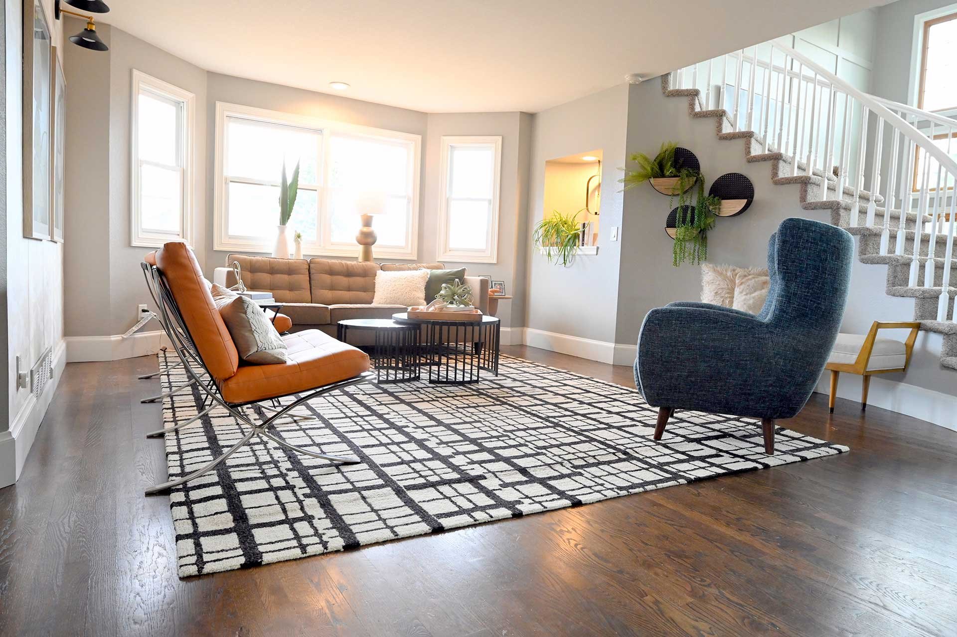 Modern residential interior design Denver living room with sunlit windows, tan sofa, orange chair, velvet blue armchair, plants, and geometric rug.