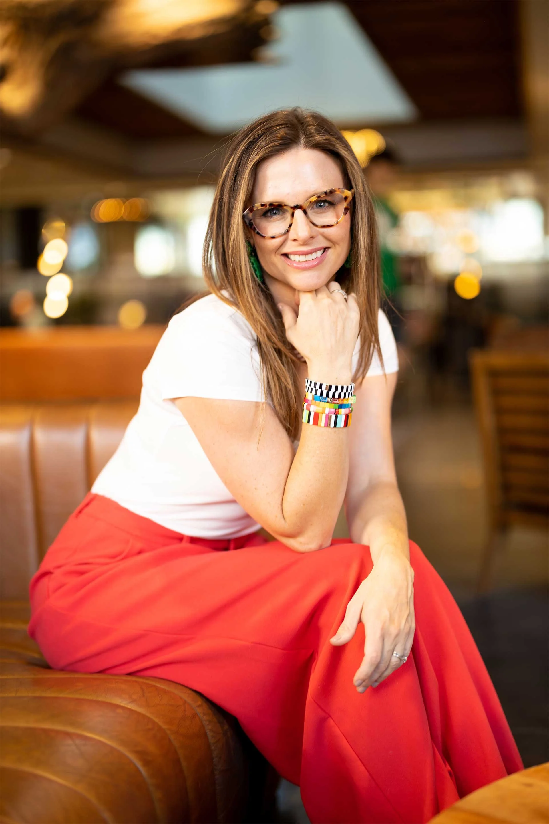 Portrait of Katie Farruggia, Interior Designer in Denver CO, wearing glasses and colorful bracelets, smiling in a warm, modern lounge..