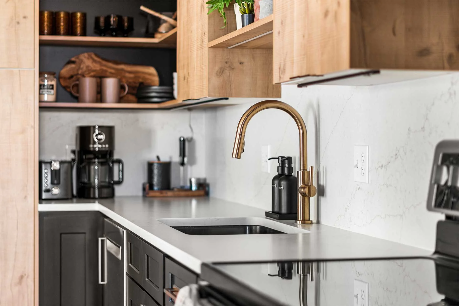 Close-up of sleek sink area with gold-toned faucet, black dispenser, and open wood cabinets designed by Make The Room in Denver, CO.