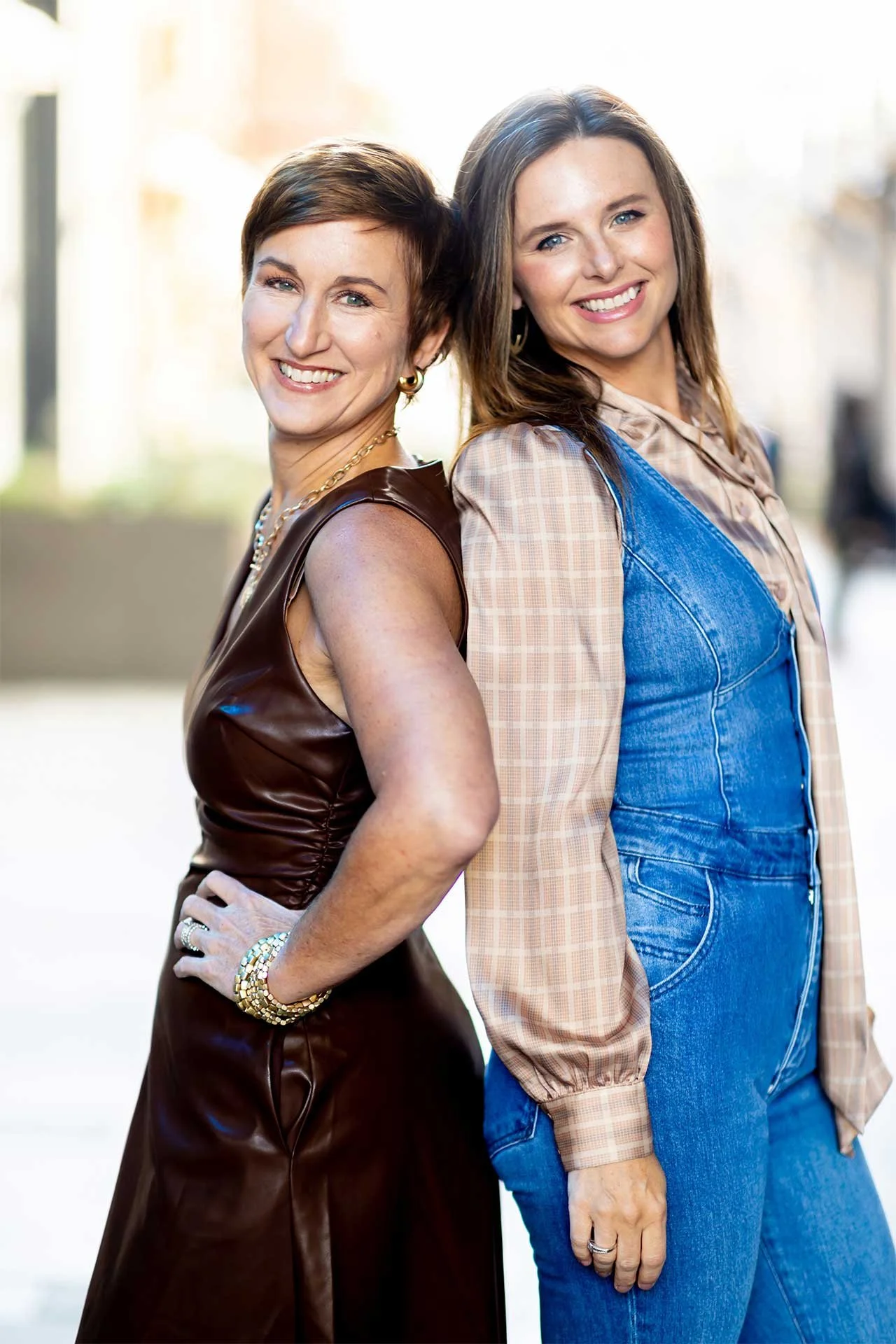 Jen and Katie, interior designers in Denver, smile together outdoors, one in brown leather dress beside the other in denim overalls.