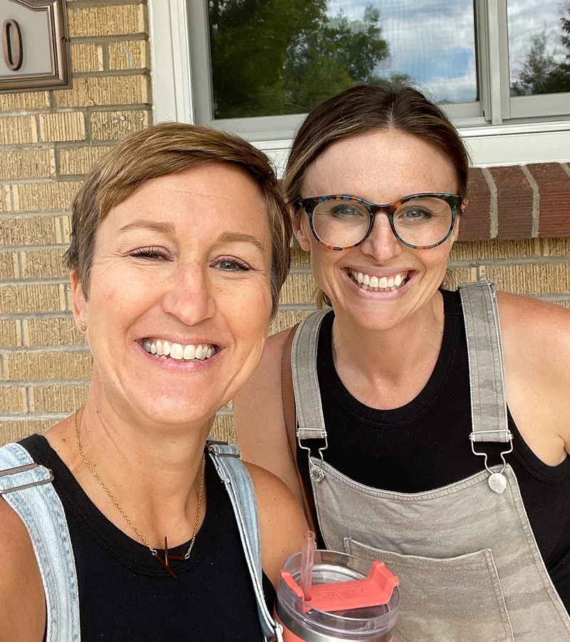 Denver interior designers Jen and Katie pose happily outdoors near a window, wearing casual tops and overalls with a drink.