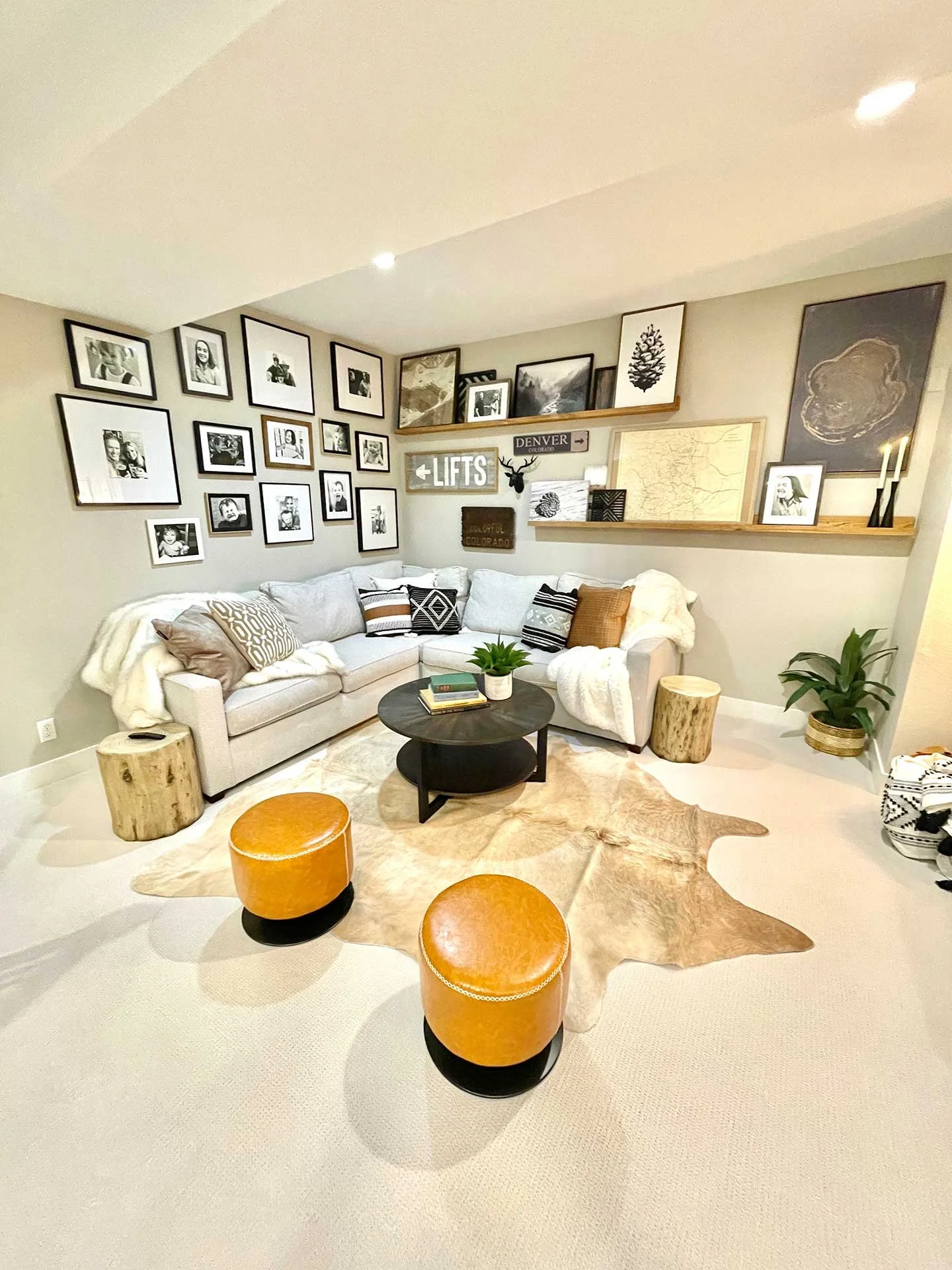 Cozy basement lounge with sectional sofa, gallery wall of framed photos and art, leather ottomans, hide rug, and wood accents with layered shelving.